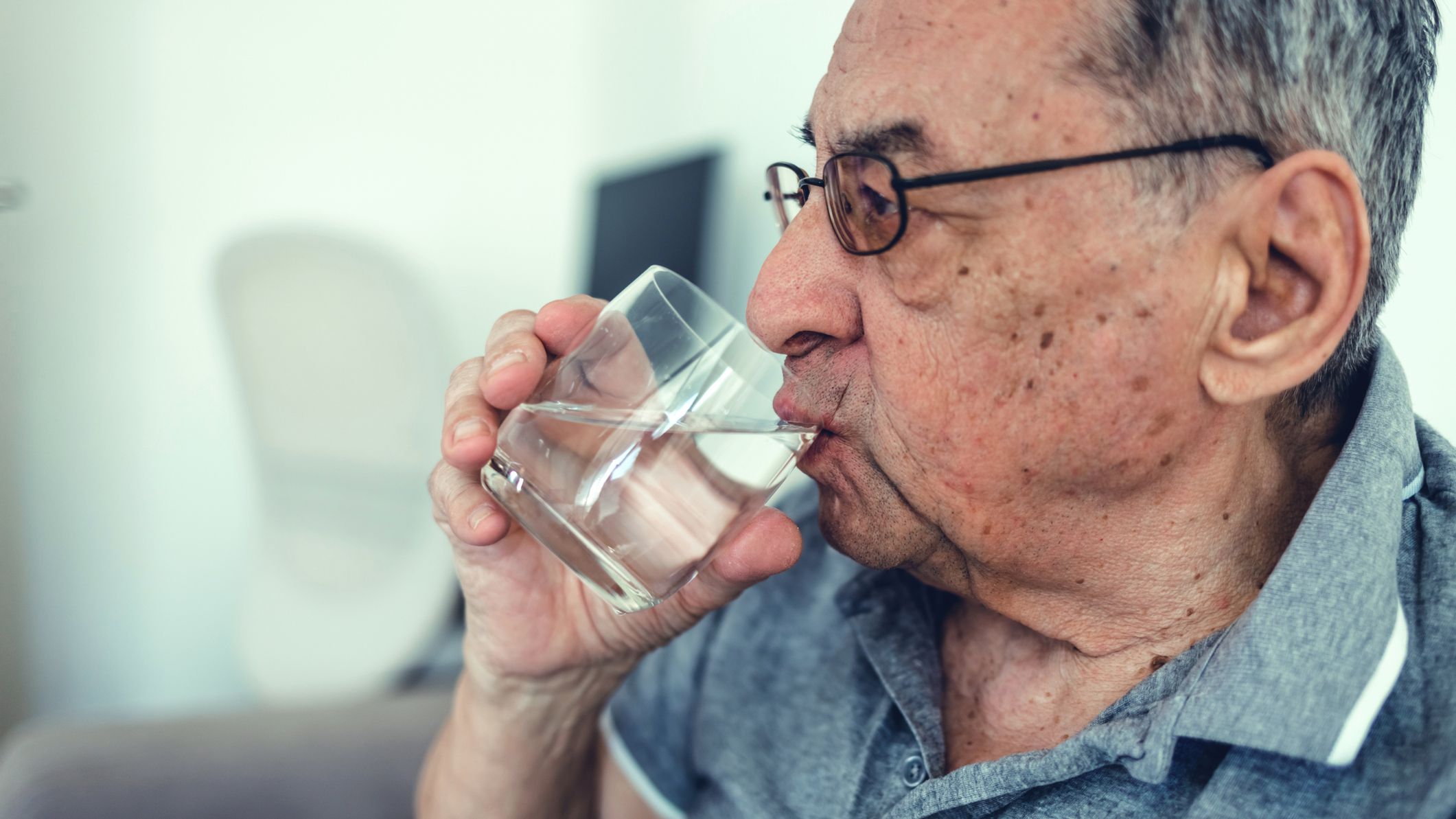 Close-up of an older person wearing glasses drinking water from a clear glass.