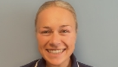 Smiling person wearing a dark blue nurse uniform standing against a plain light blue background.