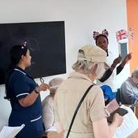 A group of people in a room, some wearing festive headbands and holding papers or small Union Jack flags.