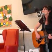 Woman singing and playing guitar indoors next to a red armchair