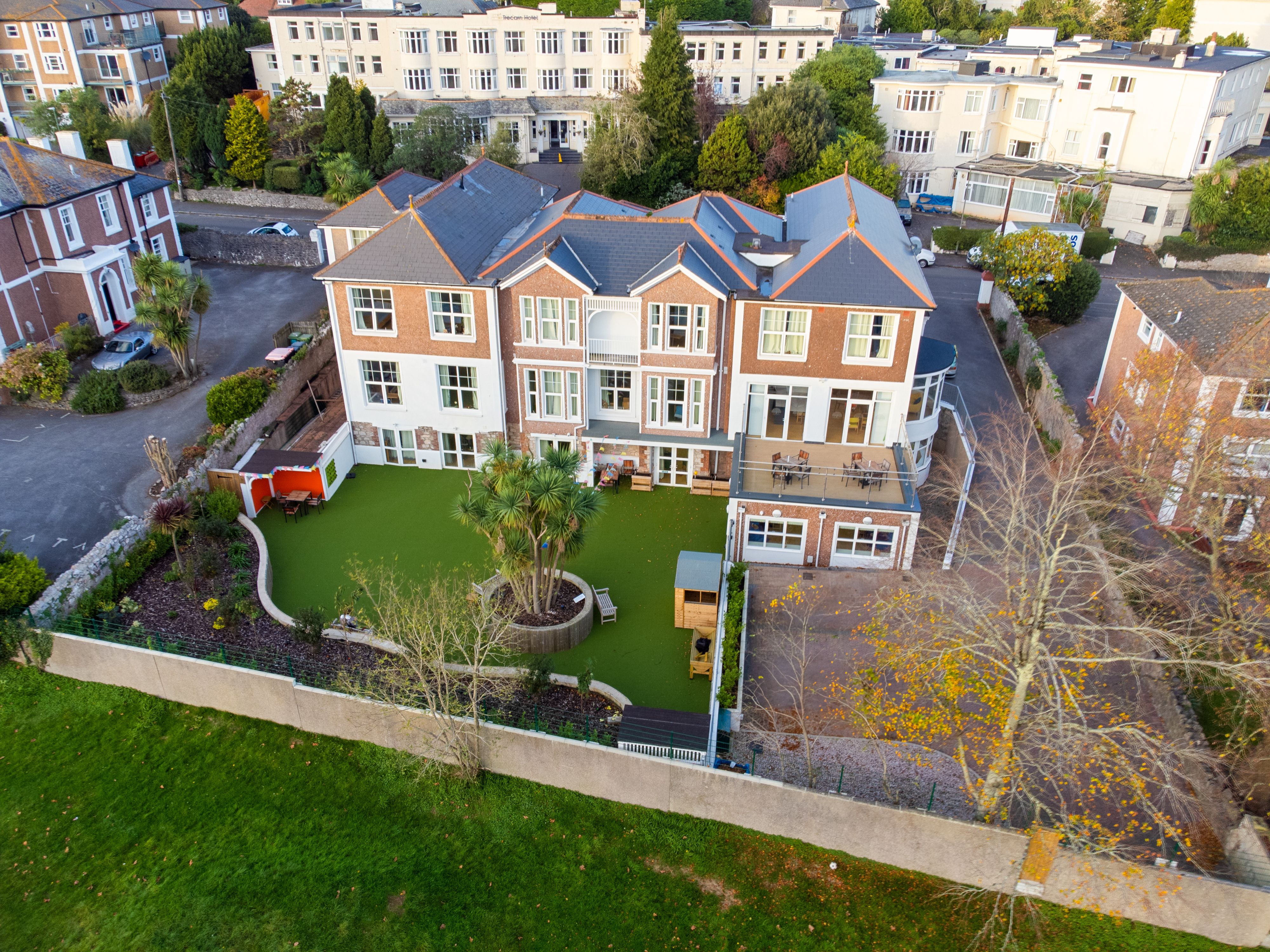 An aerial view of a large red-brick building with a neatly landscaped garden featuring artificial grass, palm trees, and seating areas, surrounded by other properties in a suburban setting.