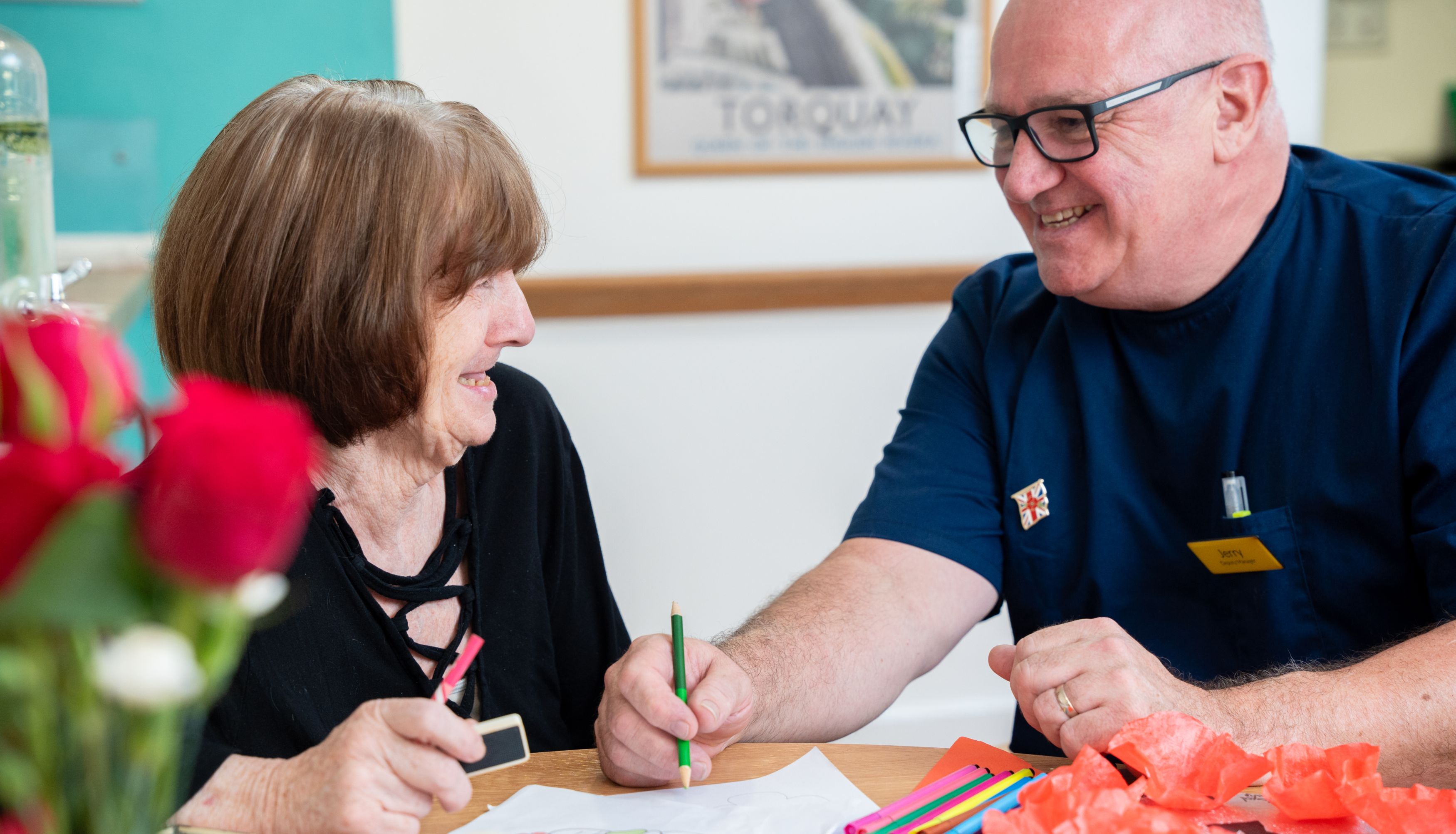 A smiling elderly woman and a man in a blue uniform sit at a table doing arts and crafts together in a care home in Torbay, surrounded by colorful materials and a vase of red roses.