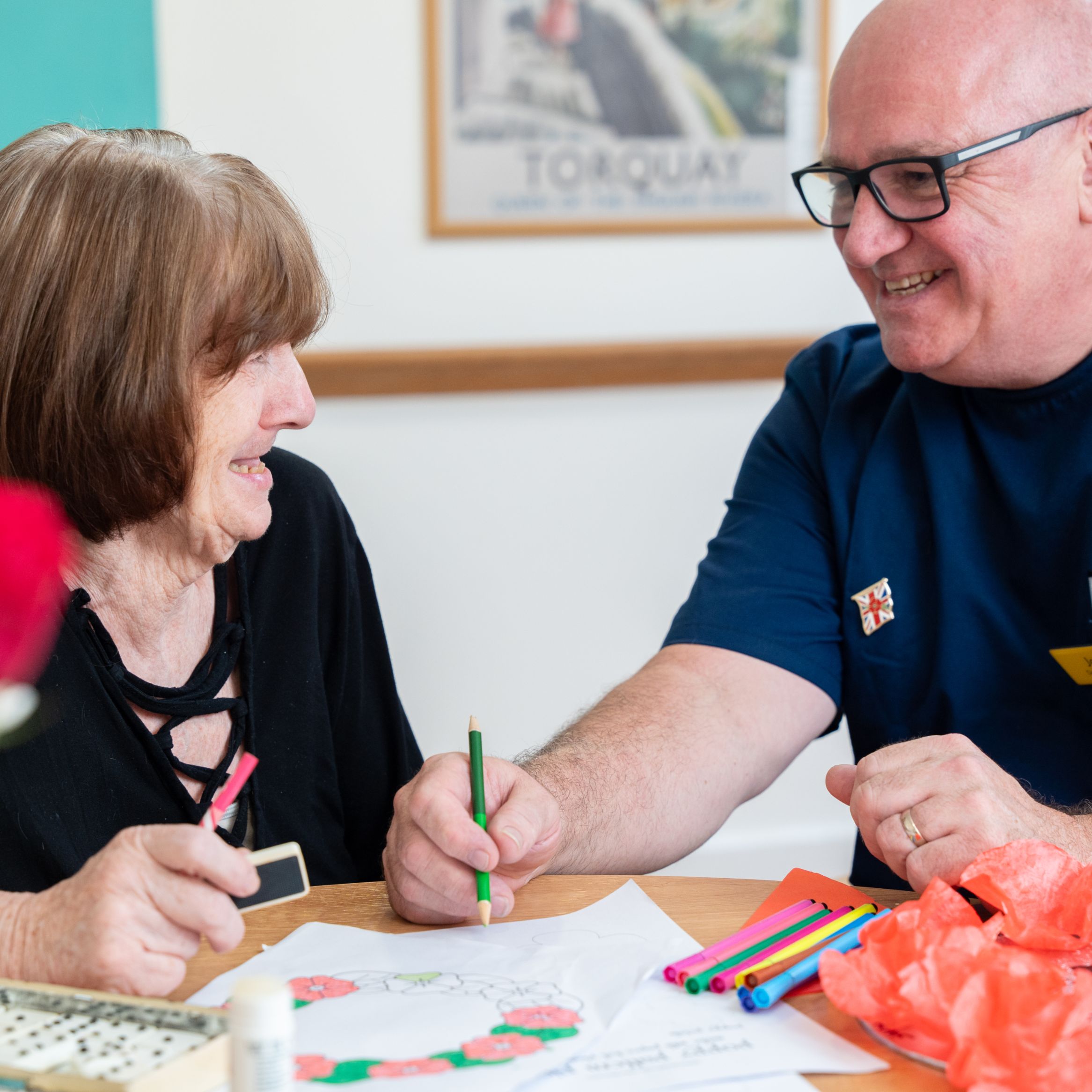 A smiling elderly woman and a man in a blue uniform sit at a table doing arts and crafts together in a care home in Torbay, surrounded by colorful materials and a vase of red roses.