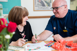A smiling elderly woman and a man in a blue uniform sit at a table doing arts and crafts together in a care home in Torbay, surrounded by colorful materials and a vase of red roses.