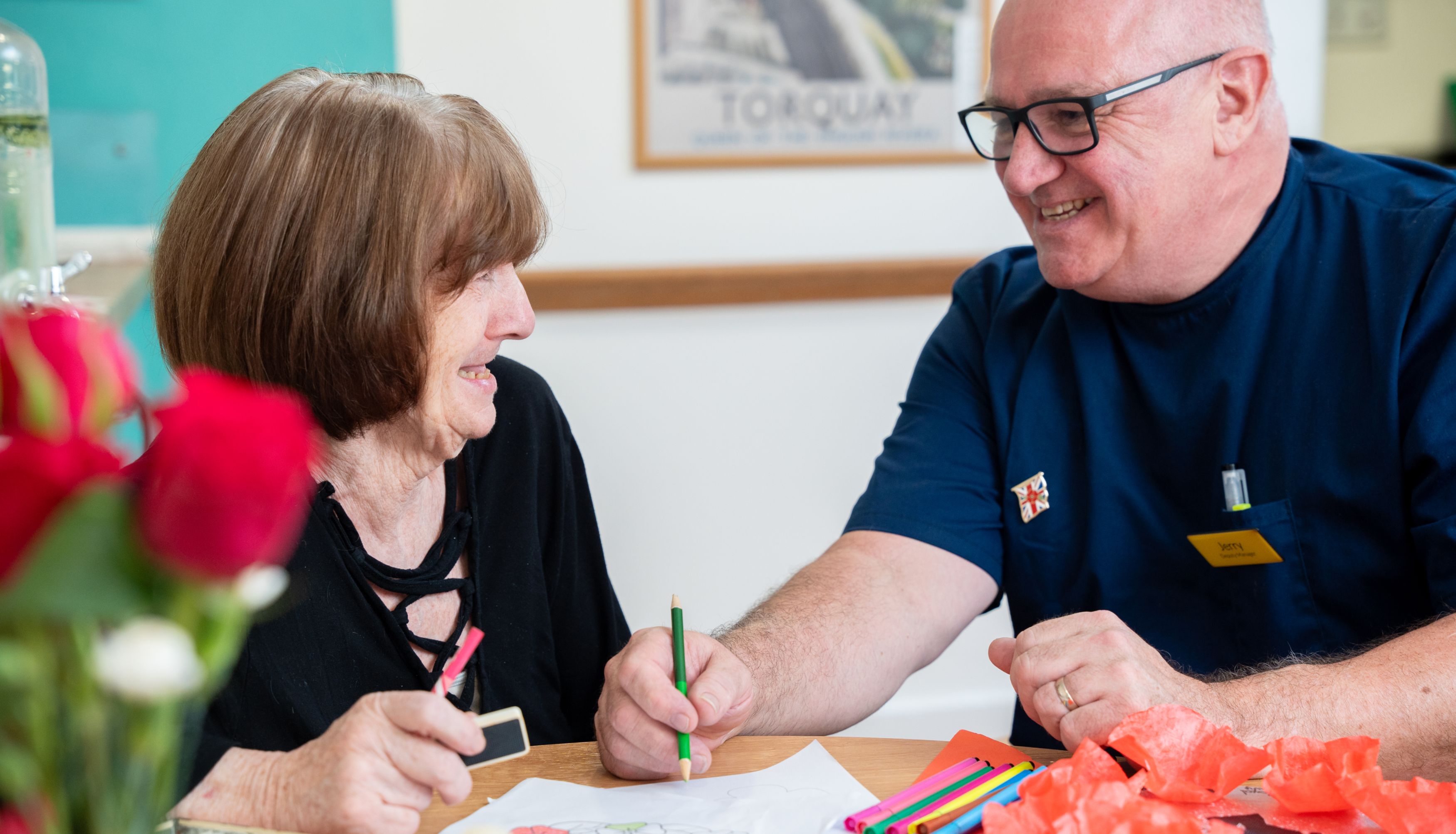 A smiling elderly woman and a man in a blue uniform sit at a table doing arts and crafts together in a care home in Torbay, surrounded by colorful materials and a vase of red roses.