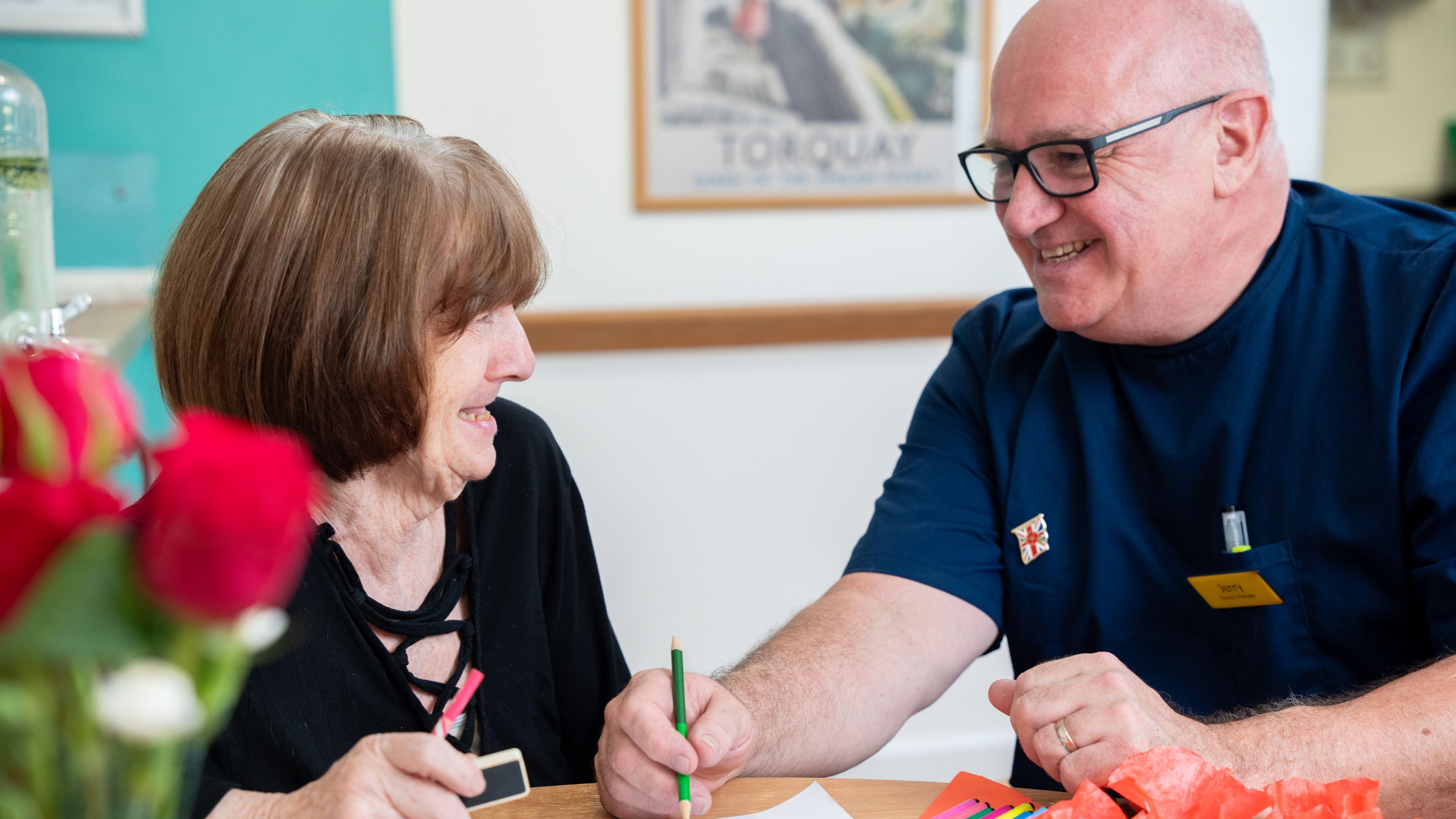 A smiling elderly woman and a man in a blue uniform sit at a table doing arts and crafts together in a care home in Torbay, surrounded by colorful materials and a vase of red roses.