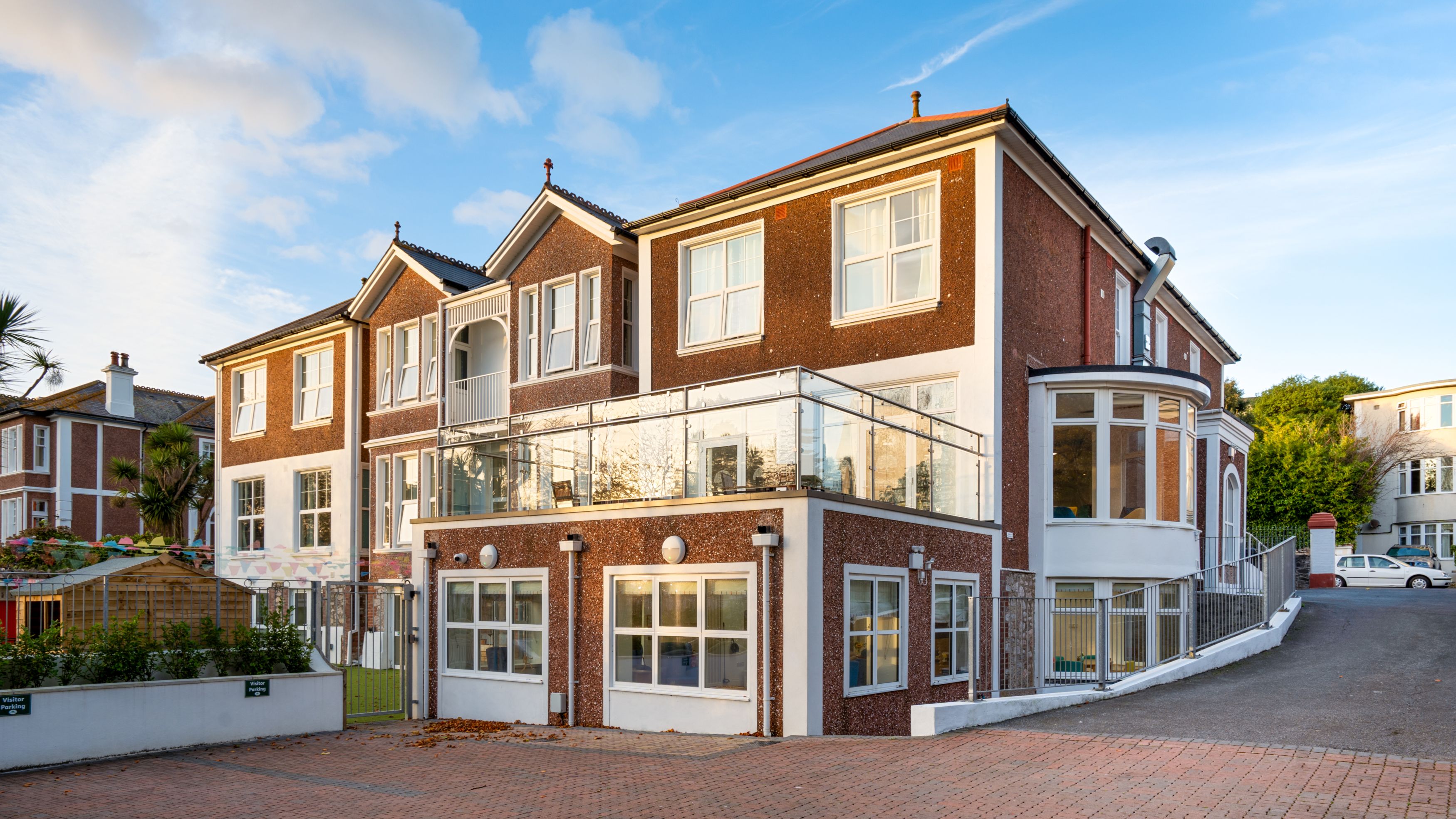 A modern red-brick building with large windows, a glass balcony, and a curved extension. The front features a paved driveway, landscaped garden, and a clear blue sky in the background.