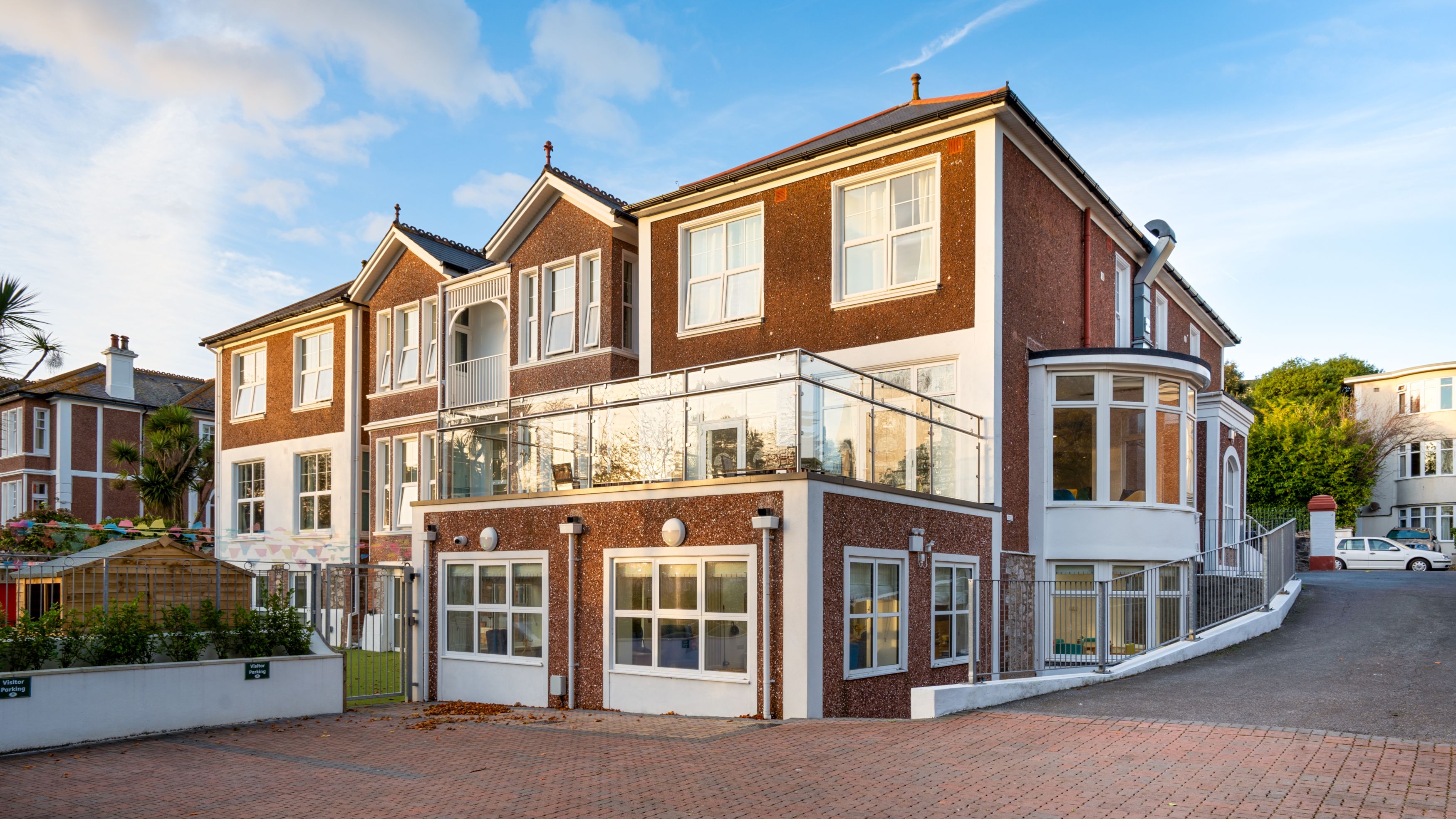 A modern red-brick building with large windows, a glass balcony, and a curved extension. The front features a paved driveway, landscaped garden, and a clear blue sky in the background.
