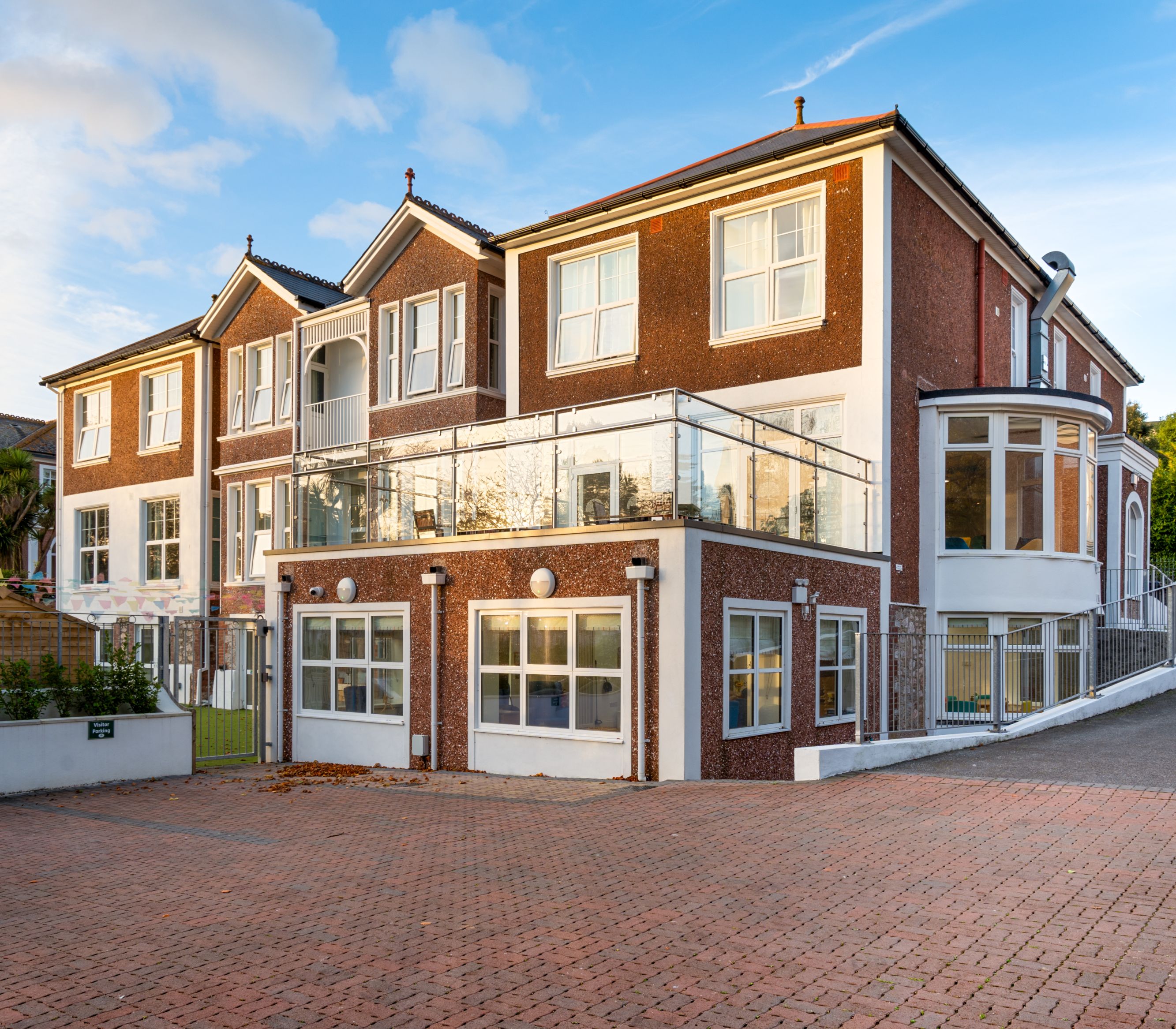 A modern red-brick building with large windows, a glass balcony, and a curved extension. The front features a paved driveway, landscaped garden, and a clear blue sky in the background.