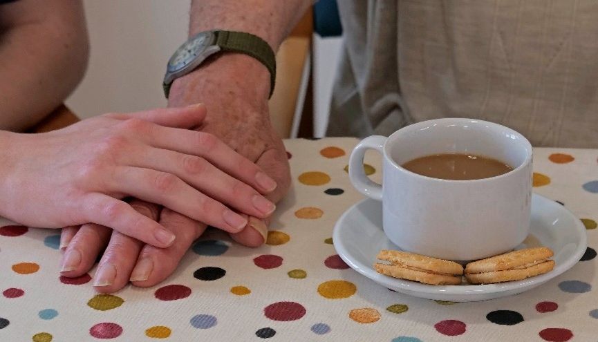 Two hands, one younger and one older, rest gently together on a polka-dot tablecloth next to a white cup of tea and three biscuits on a saucer, creating a warm and comforting scene.