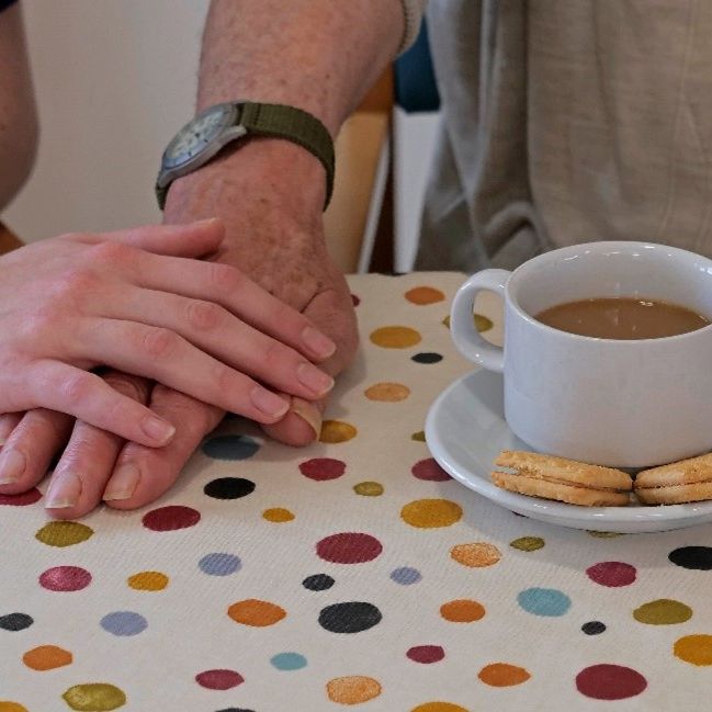 Two hands, one younger and one older, rest gently together on a polka-dot tablecloth next to a white cup of tea and three biscuits on a saucer, creating a warm and comforting scene.