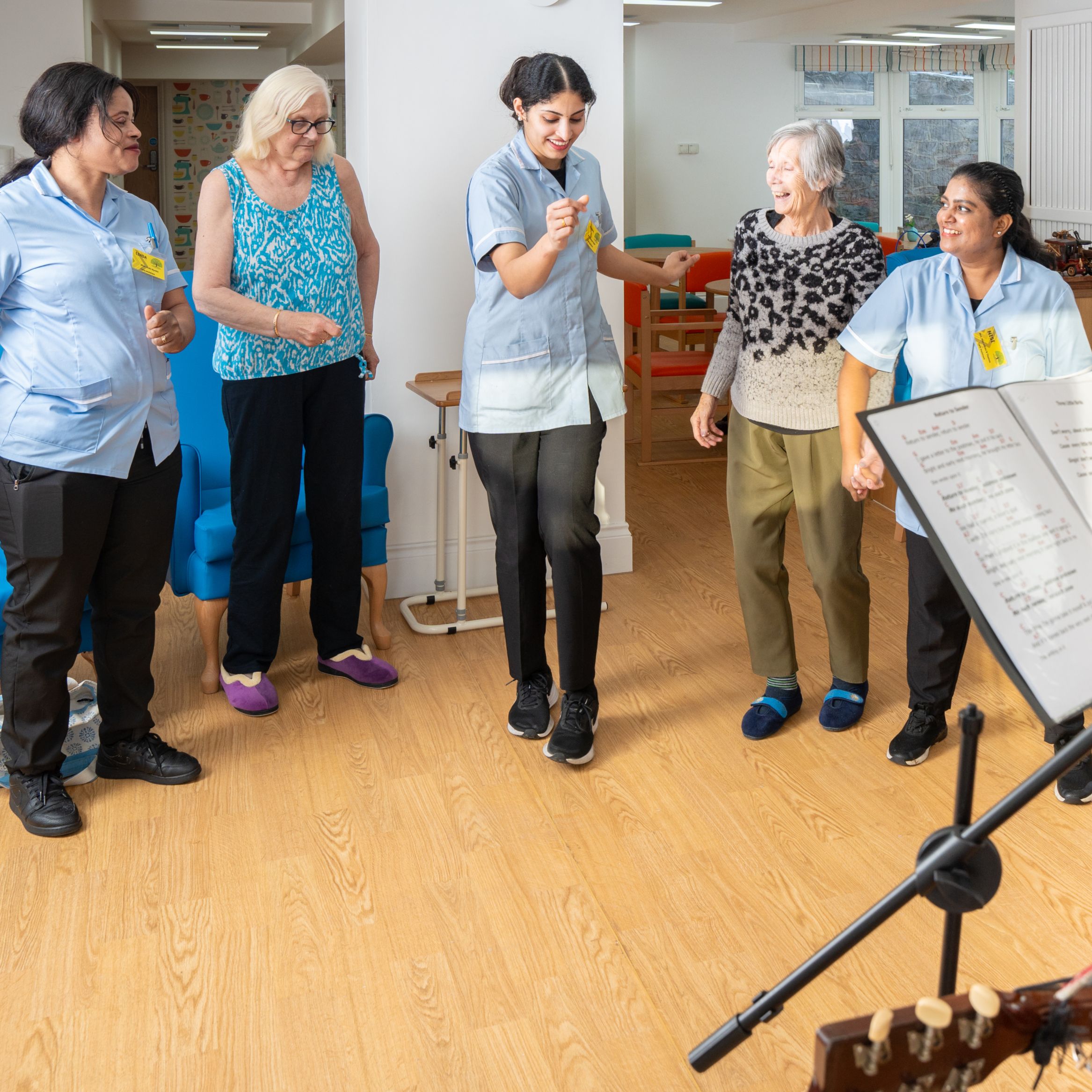 Care home staff and residents dance joyfully during a musical performance as a guitarist and singer play. An elderly woman observes from a blue armchair in the cheerful room.