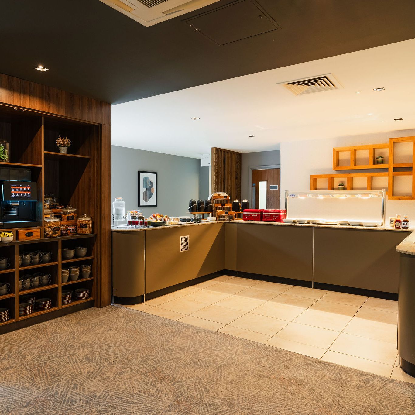 Modern hotel breakfast buffet area with beverage dispensers, food display, and shelves of cups and plates