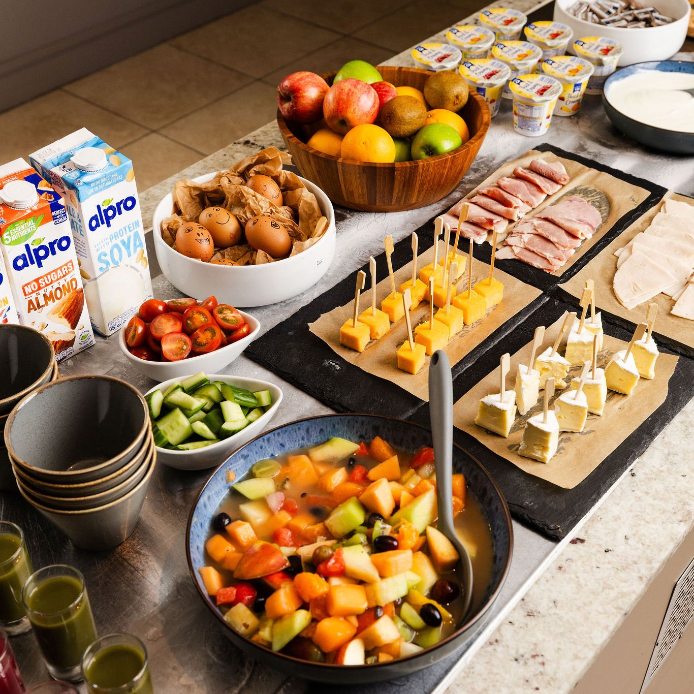 Buffet table with various breakfast options including fruit, cheeses, cold meats, plant-based milks, and vegetable bowls.