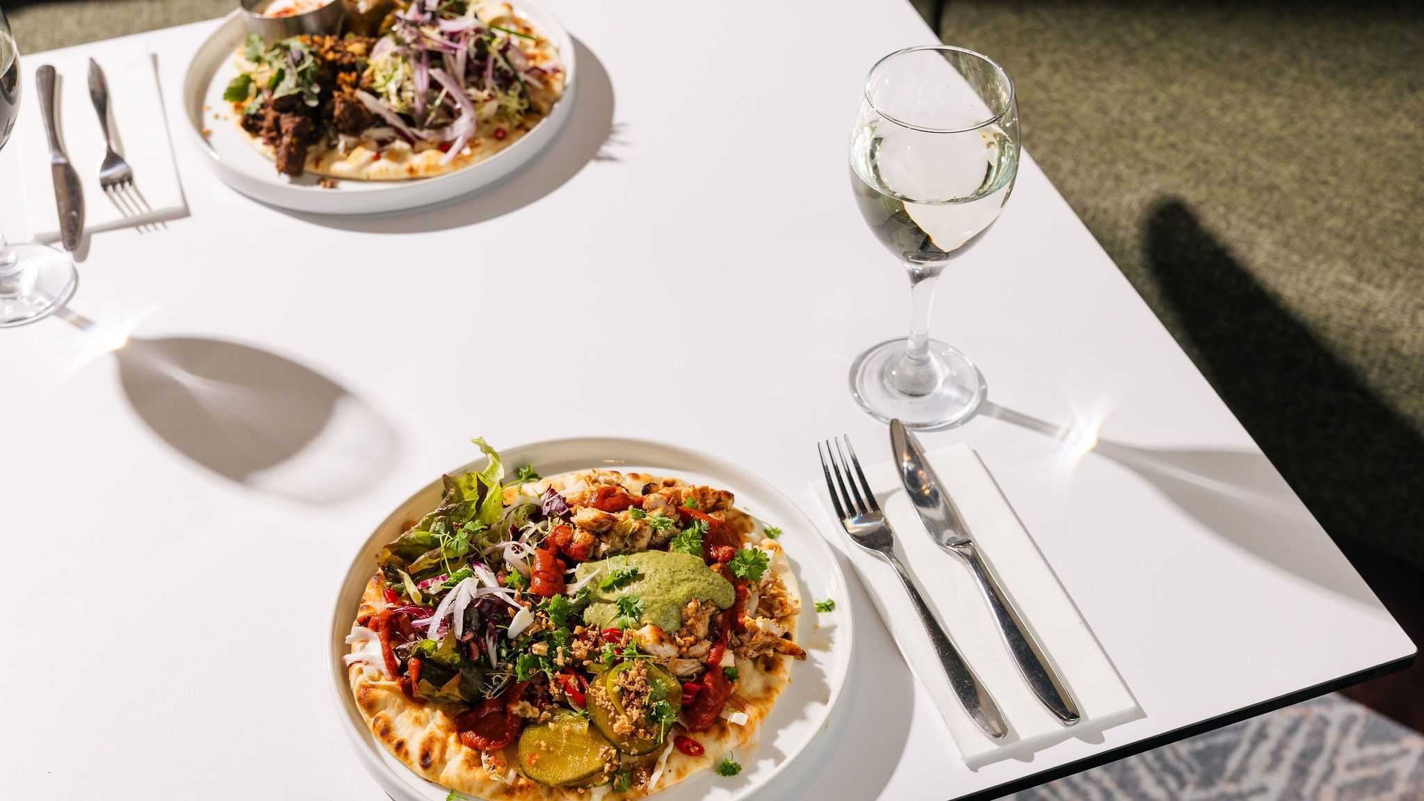 Two plates of flatbread topped with various vegetables and meat, accompanied by a glass of white wine on a white table.