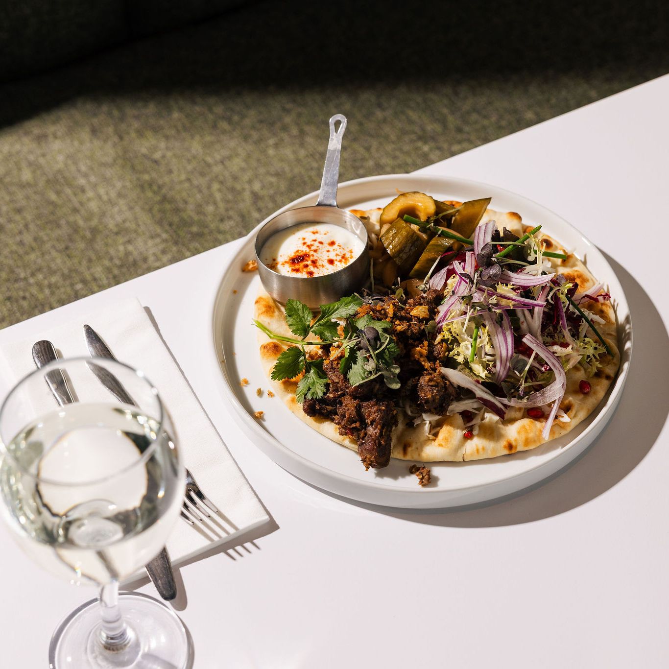 Plate of Middle Eastern-inspired food with flatbread, salad, pickled vegetables, yogurt sauce, and a glass of white wine on a white table.