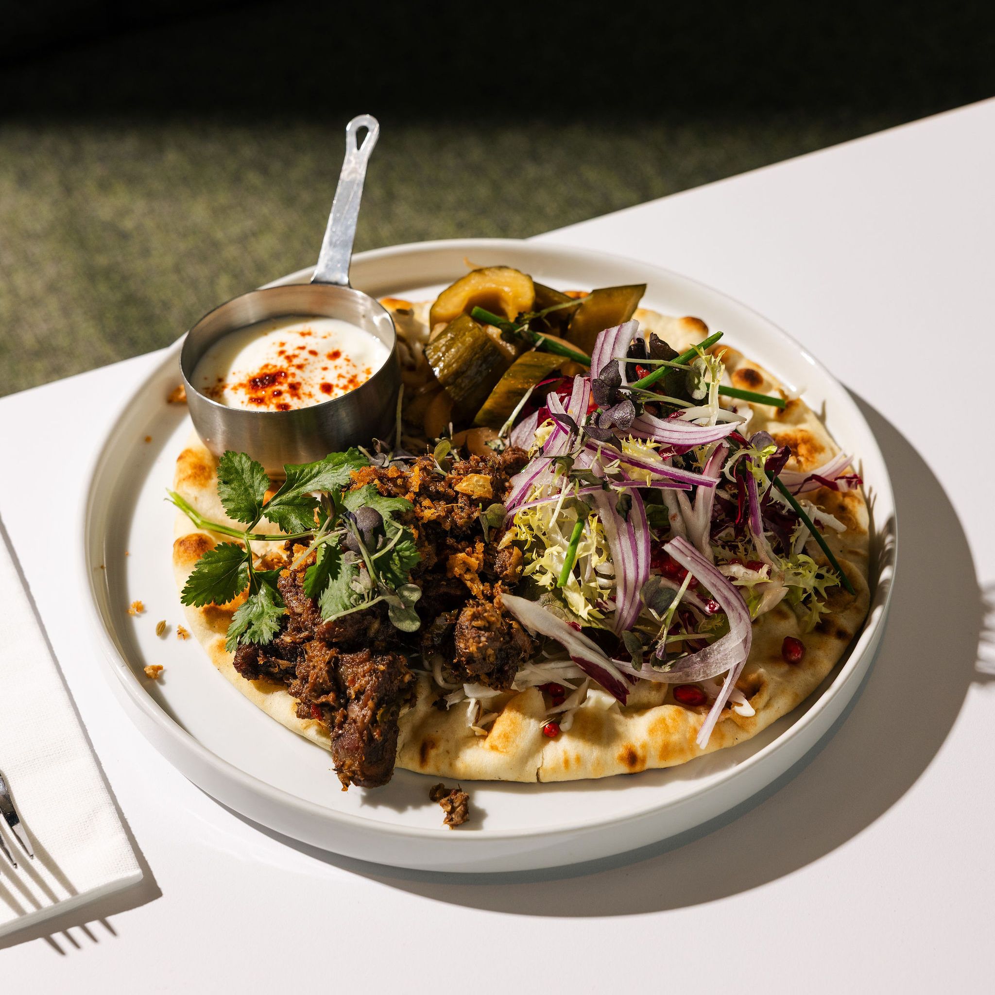 A plate of flatbread topped with marinated meat, fresh herbs, sliced onions, and pickled vegetables, served with a side of creamy white sauce.