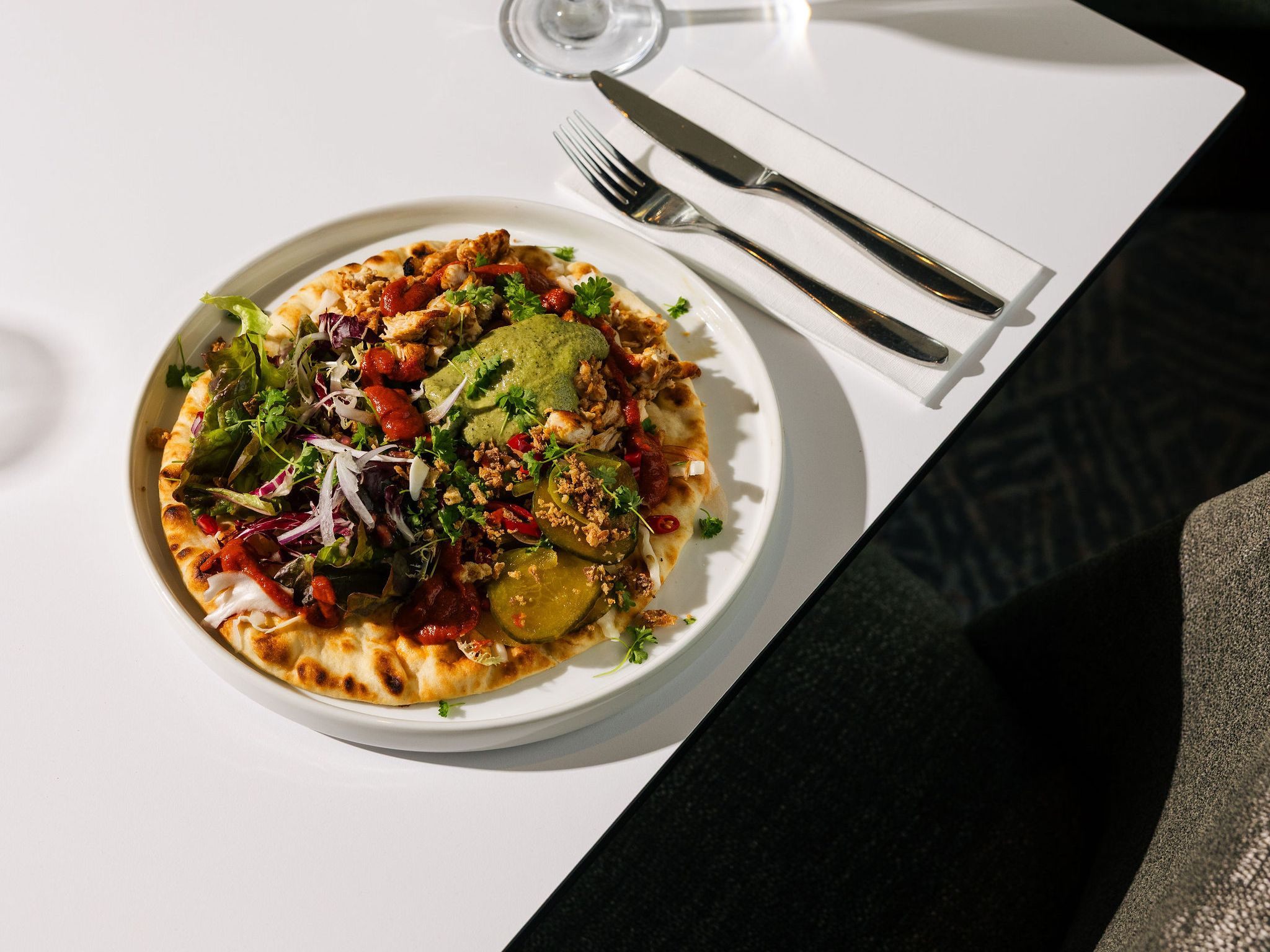 Plate of flatbread with assorted vegetables, greens, and sauce served with a glass of white wine