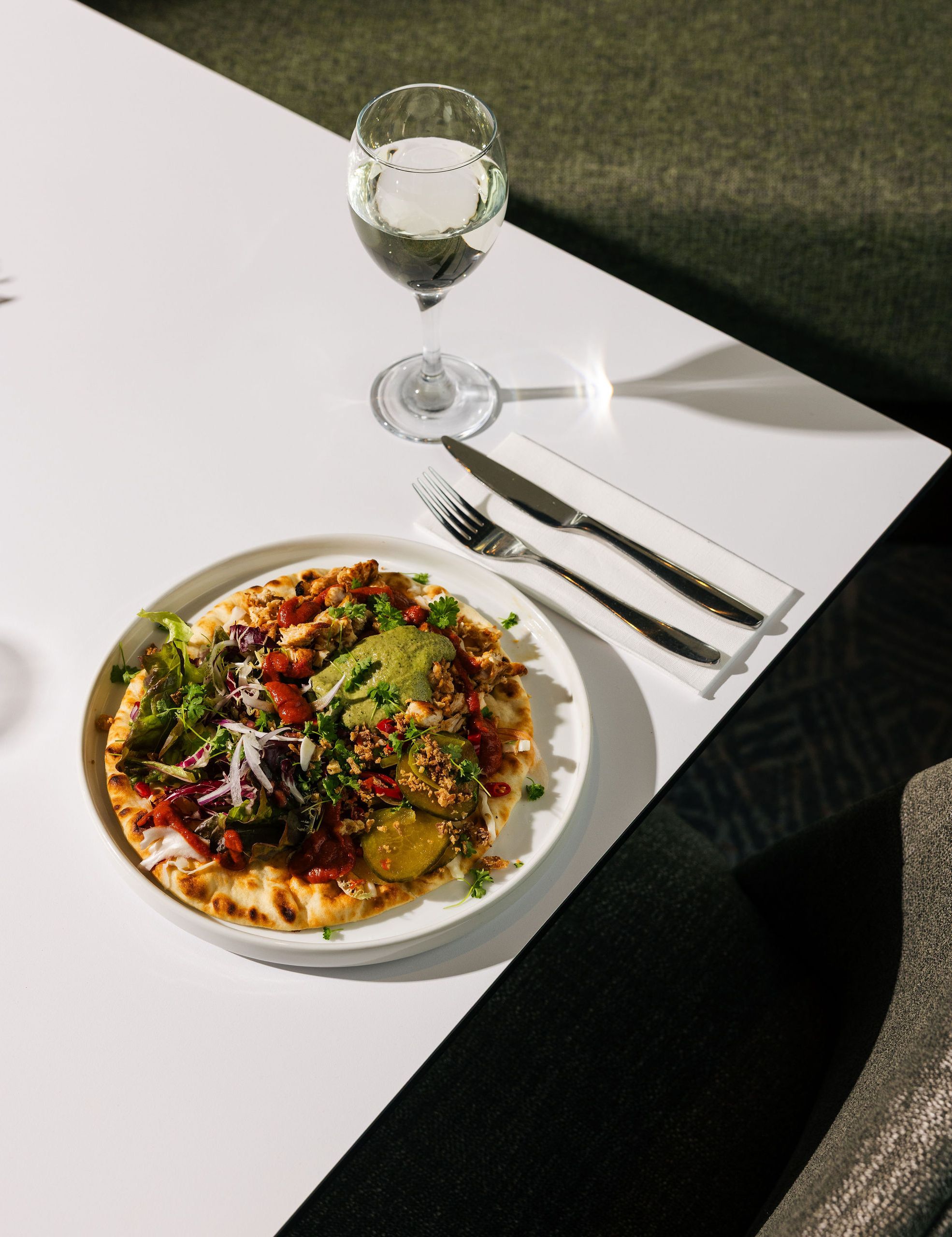 Plate of flatbread with assorted vegetables, greens, and sauce served with a glass of white wine