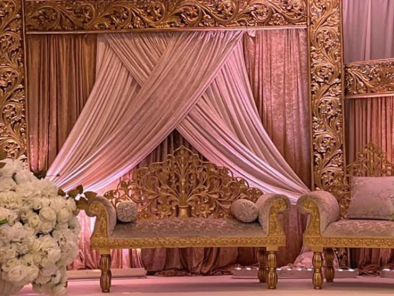 Lavishly decorated wedding stage with ornate gold furniture and draped pink curtains.