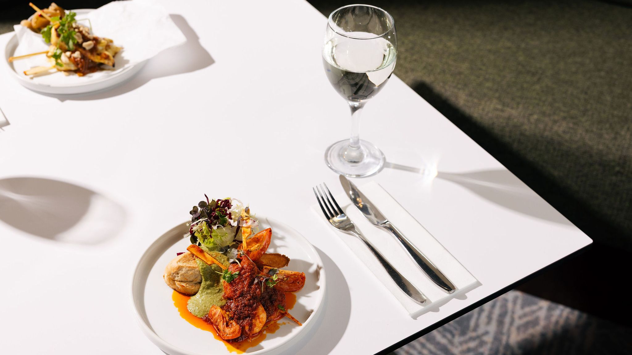 Elegant plated meal with shrimp, greens, and sauce, accompanied by a glass of white wine on a white table.