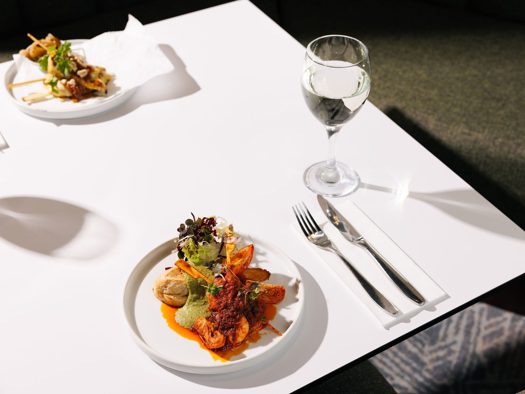 Elegant plated meal with shrimp, greens, and sauce, accompanied by a glass of white wine on a white table.