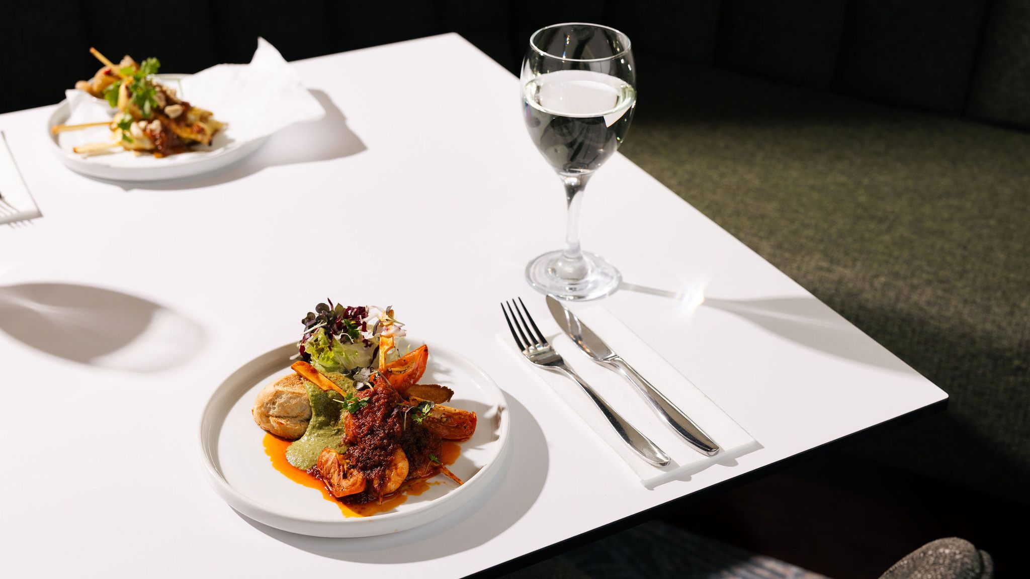 A white table with a plate of gourmet food, a glass of water, silverware, and another plate with skewers in the background.