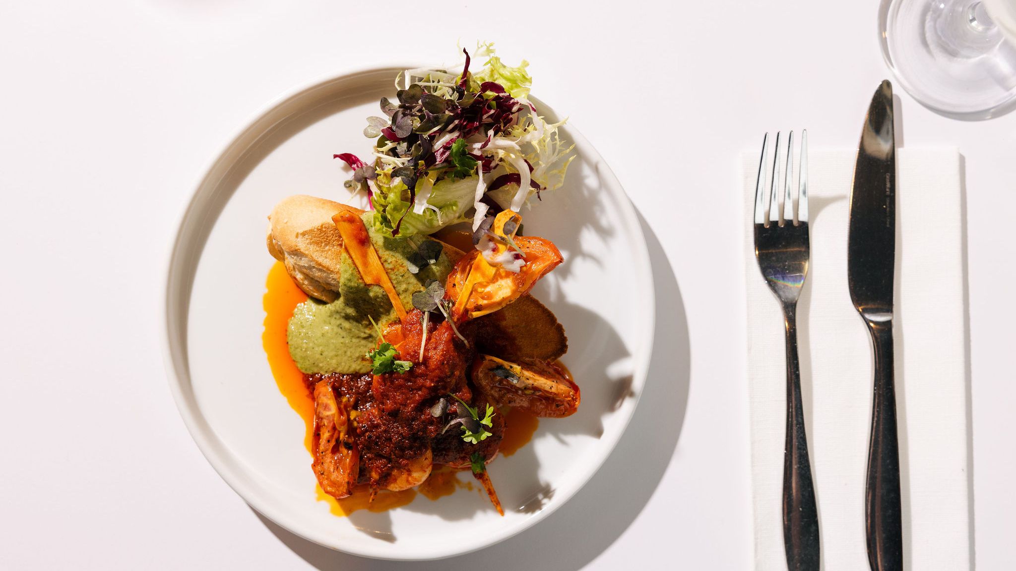 Gourmet seafood dish with prawns, bread, salad greens, and sauce on a white plate with fork and knife on a napkin.