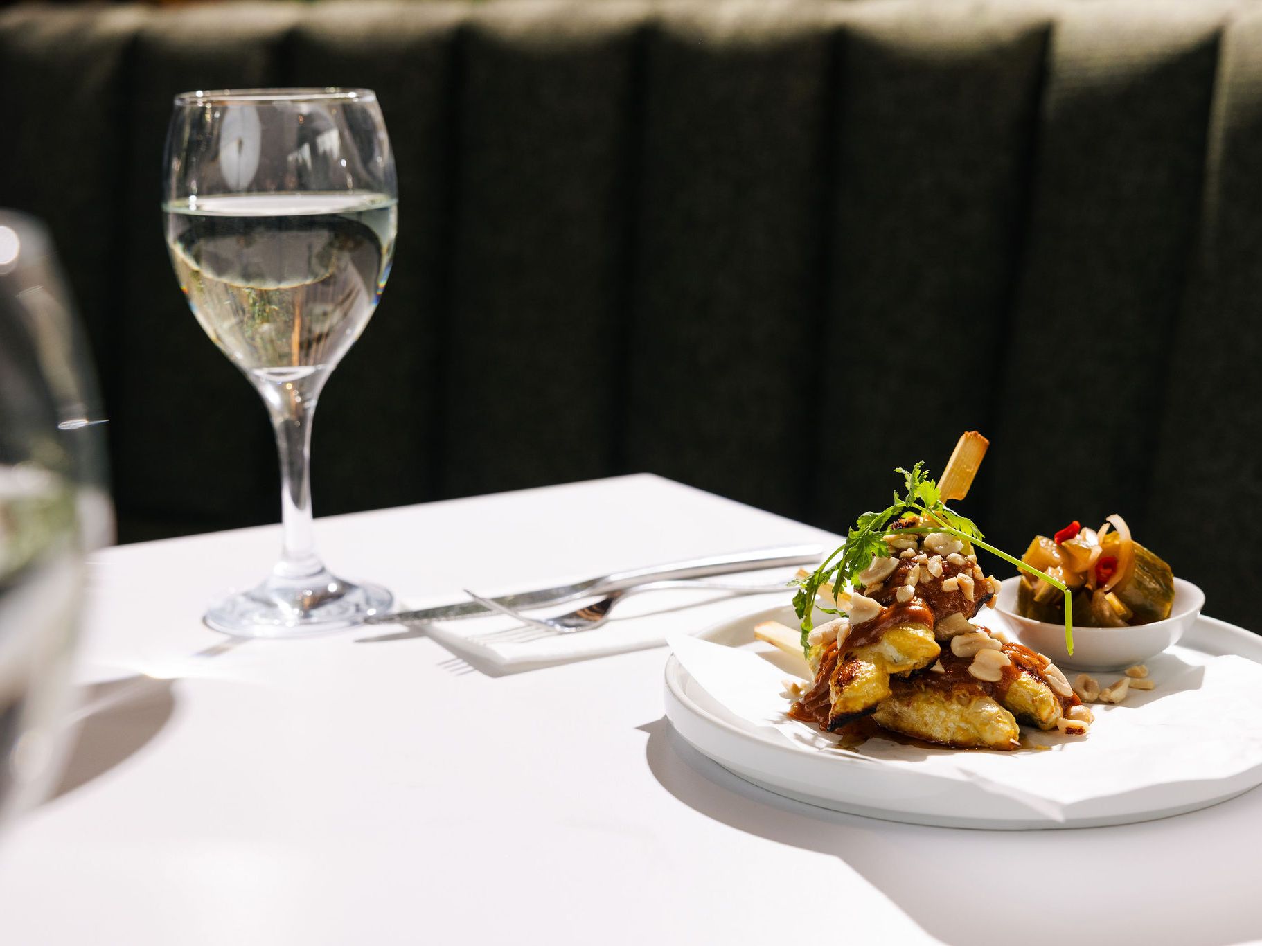 A plate of gourmet food with garnish and a side dish, next to a glass of white wine on a white table.