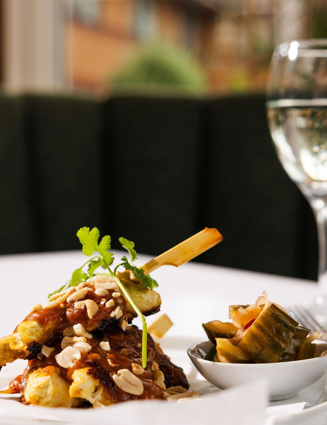 Plate of grilled skewers topped with sauce and nuts, served with pickled vegetables and a glass of white wine.