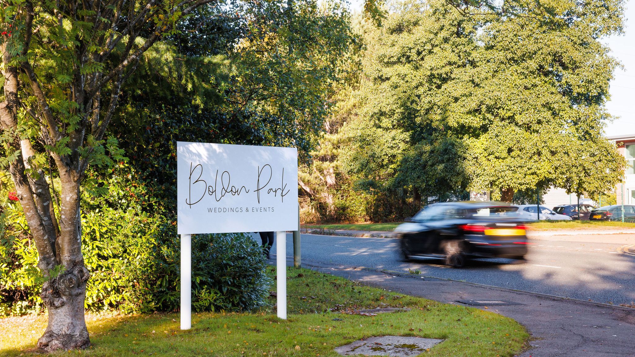 Sign for Boldon Park Weddings & Events near a road with a passing car and trees.