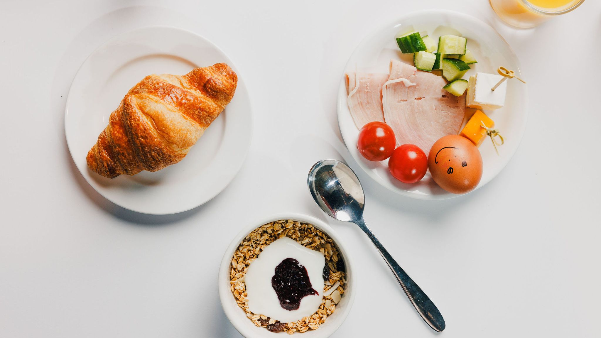 Continental breakfast with croissant, yogurt with granola and jam, cold cuts, cheese, vegetables, an egg with a face drawn on it, and orange juice.