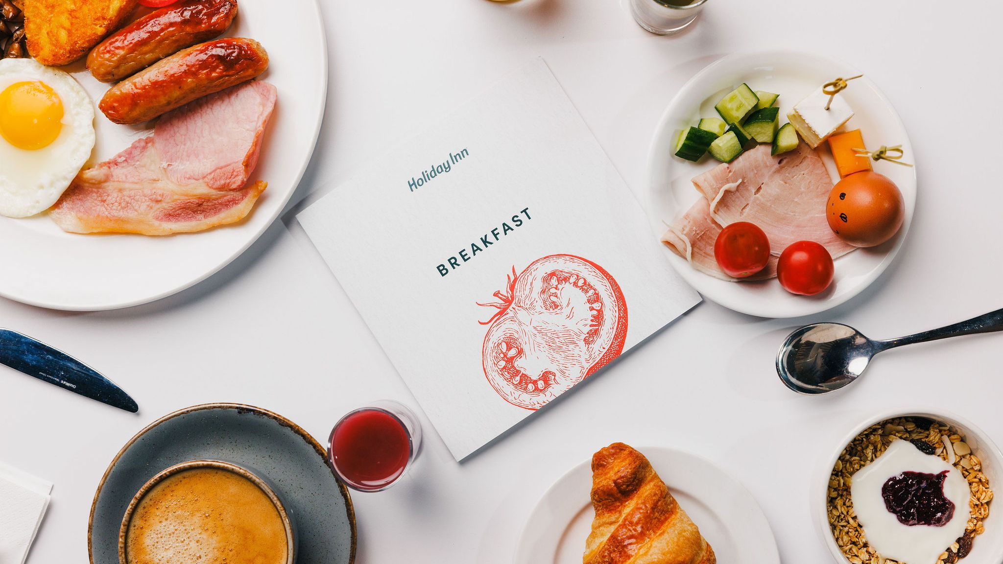 Breakfast spread with eggs, sausage, bacon, hash brown, coffee, croissant, granola with yogurt, and fresh vegetables around a Holiday Inn breakfast menu.