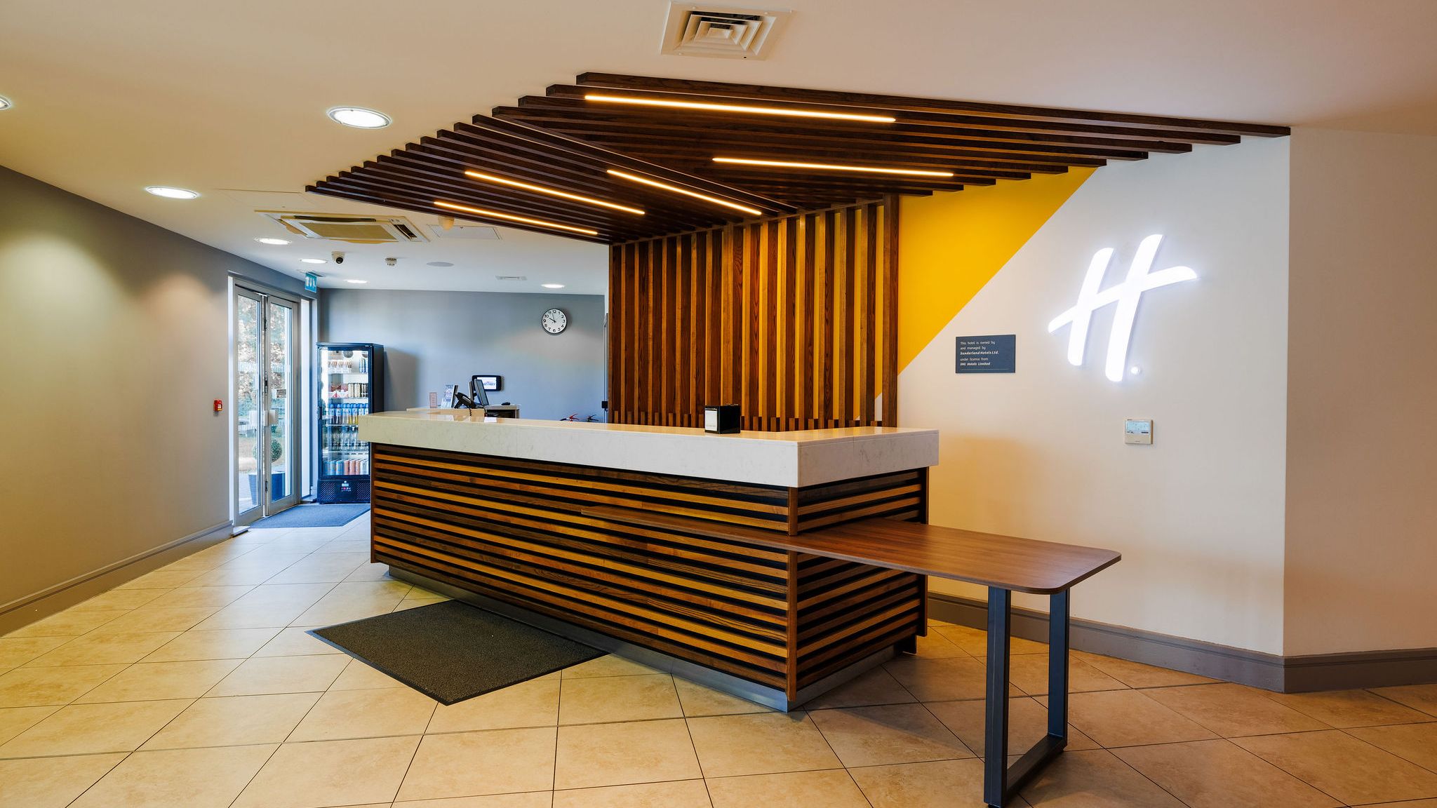 Modern hotel reception desk with wood paneling, yellow accent wall, and a white illuminated 'H' logo.