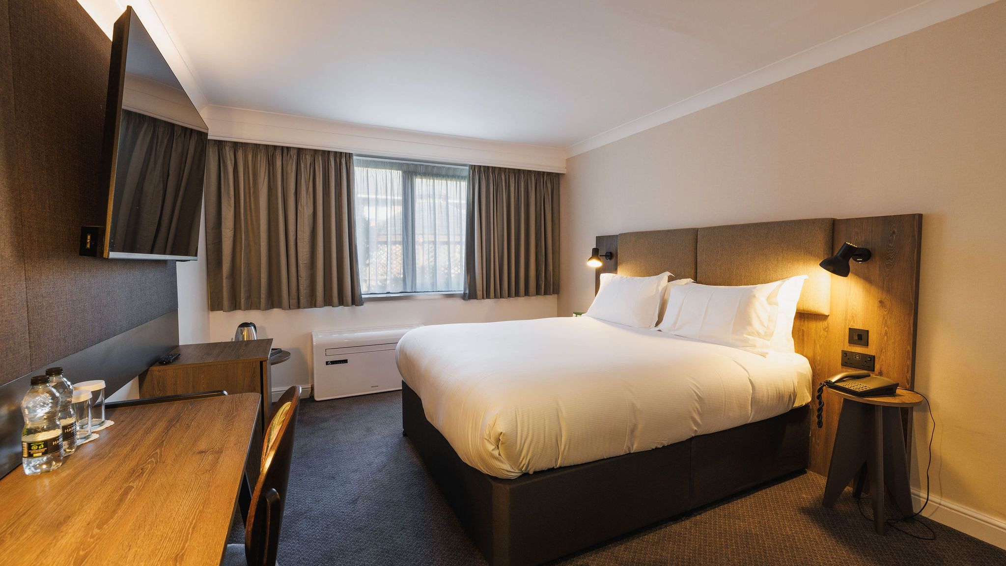 Modern hotel room with a large bed, desk, flat-screen TV, and window with curtains