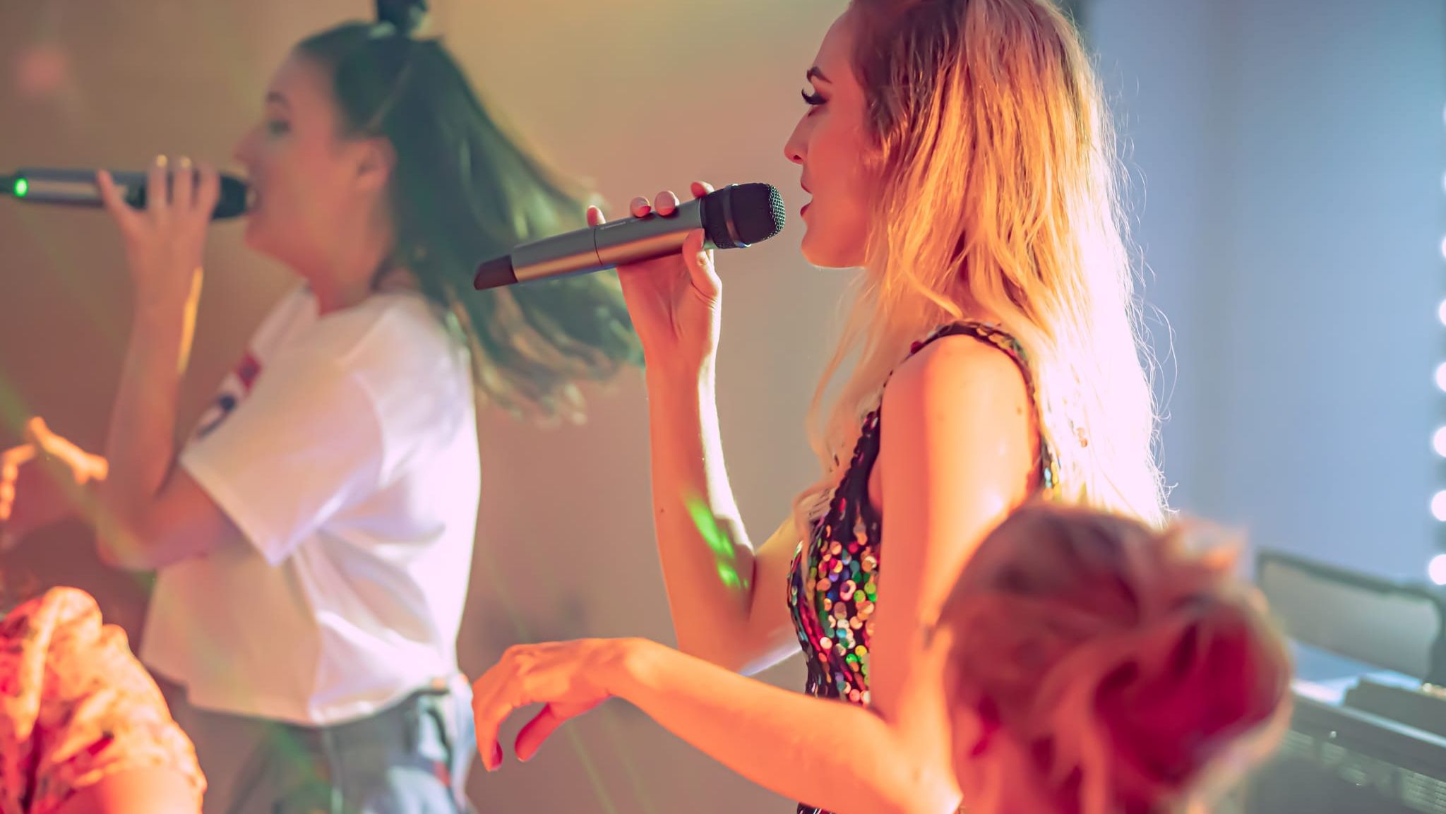 Two women singing into microphones on stage with colorful lighting