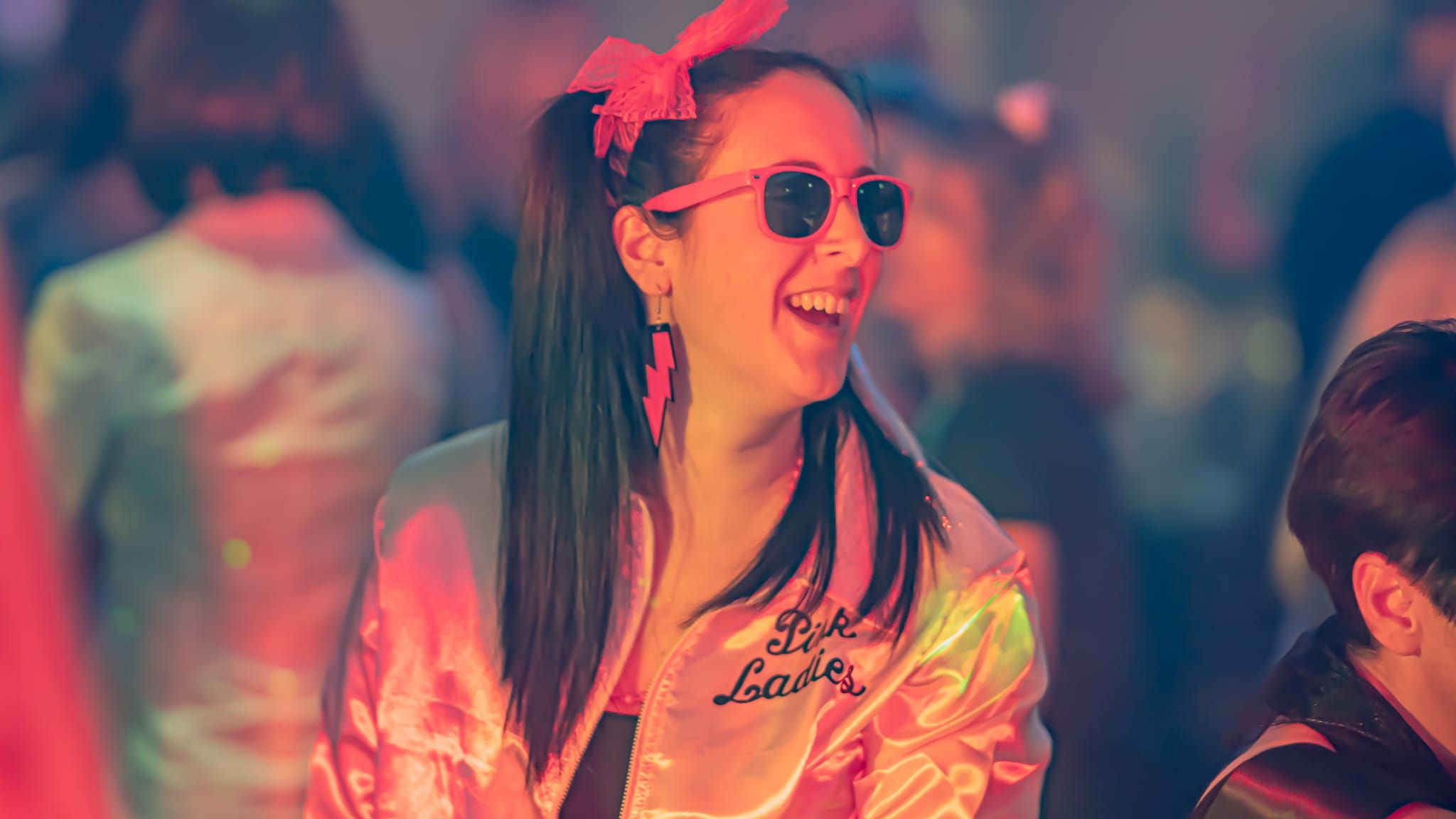 Woman wearing a pink retro jacket, sunglasses, and a bow in her hair, laughing at a party with colorful lighting