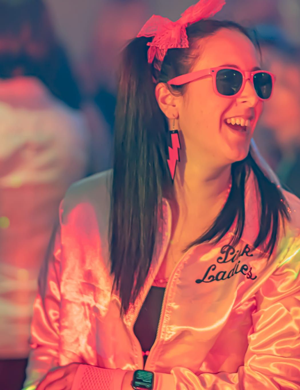 Woman wearing a pink retro jacket, sunglasses, and a bow in her hair, laughing at a party with colorful lighting