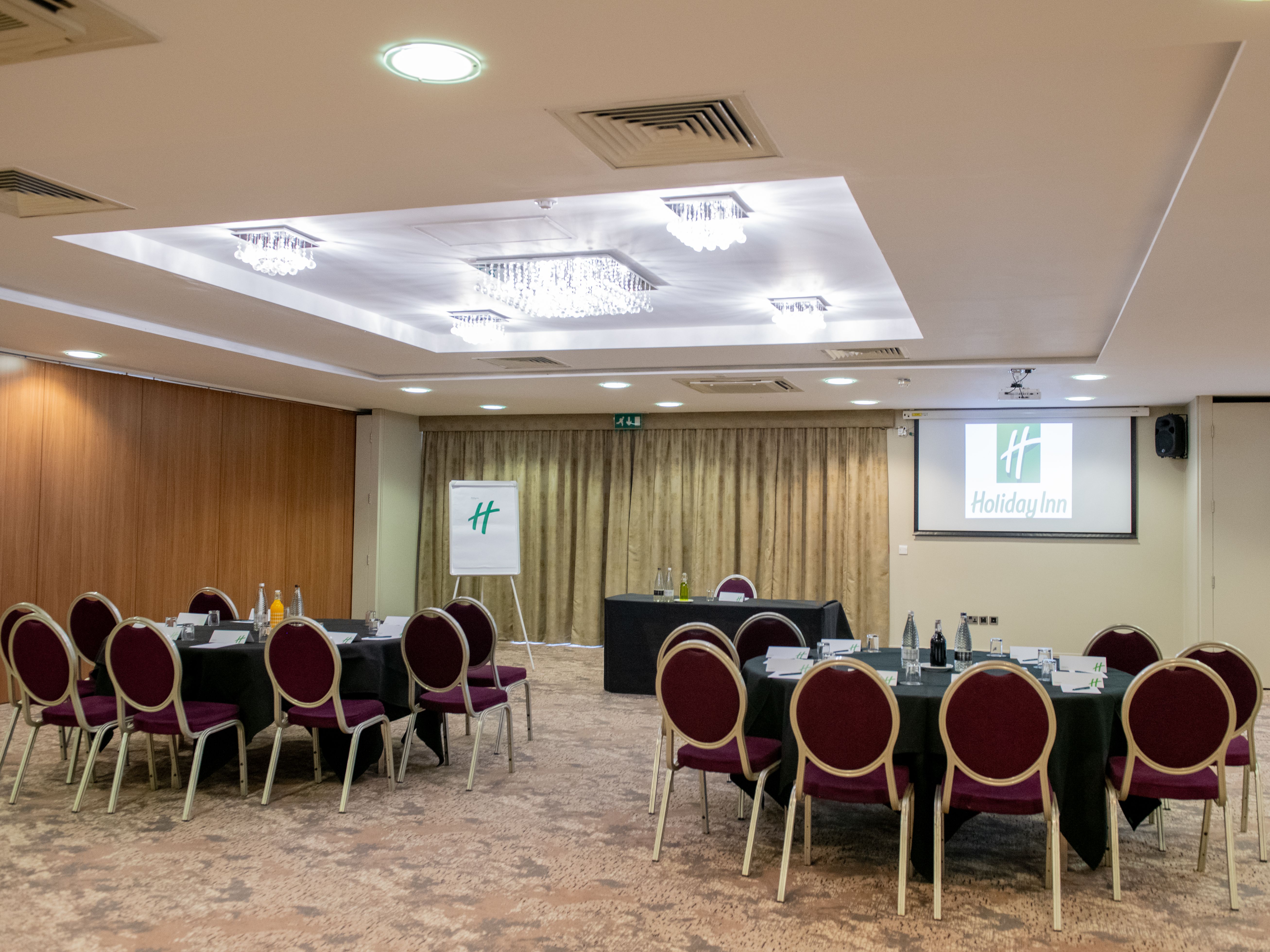 Conference room at Holiday Inn set up for a meeting with round tables and chairs, a flipchart, and a projector screen.