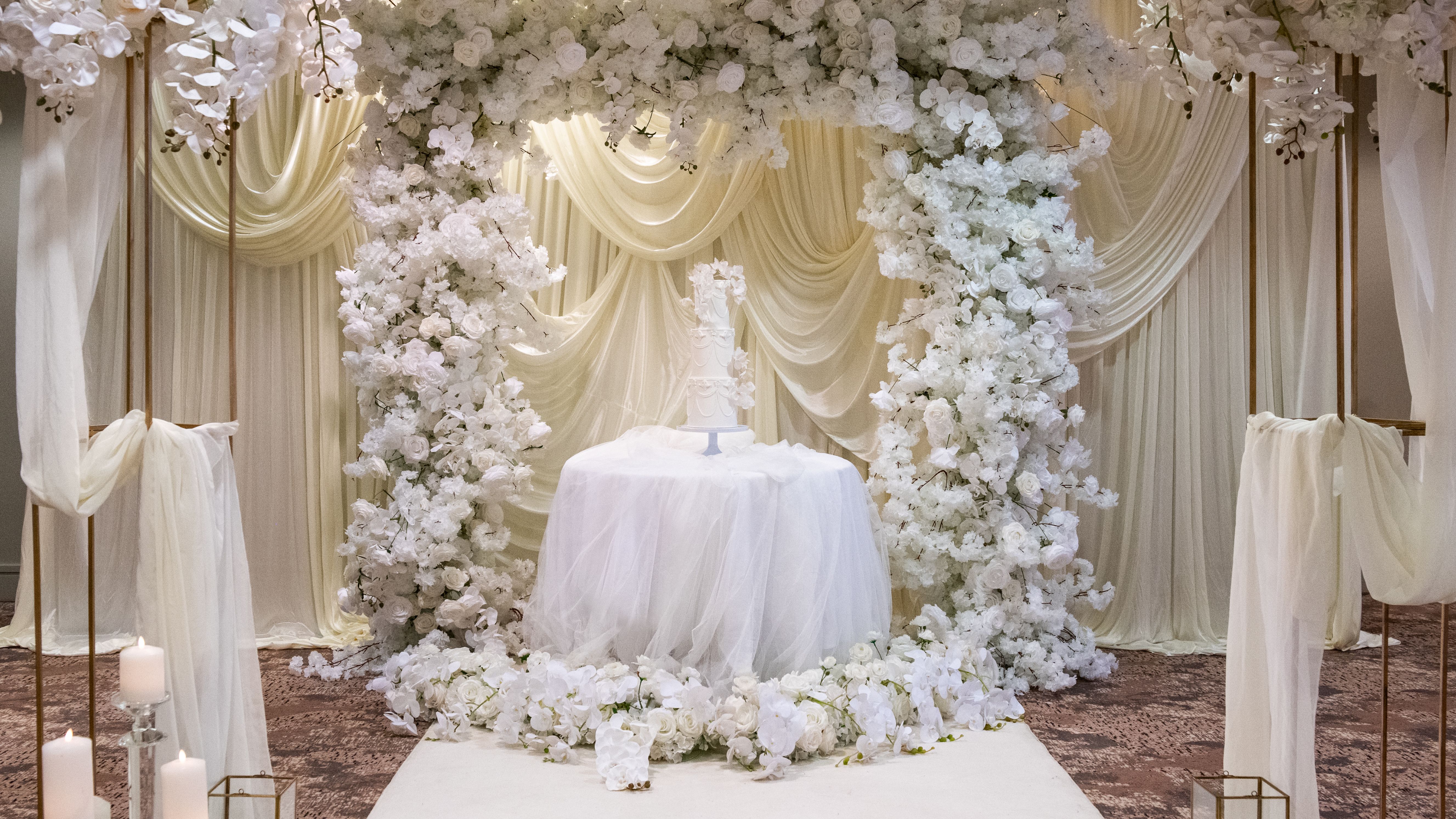 Elegant wedding cake display with white floral arch and draped backdrop