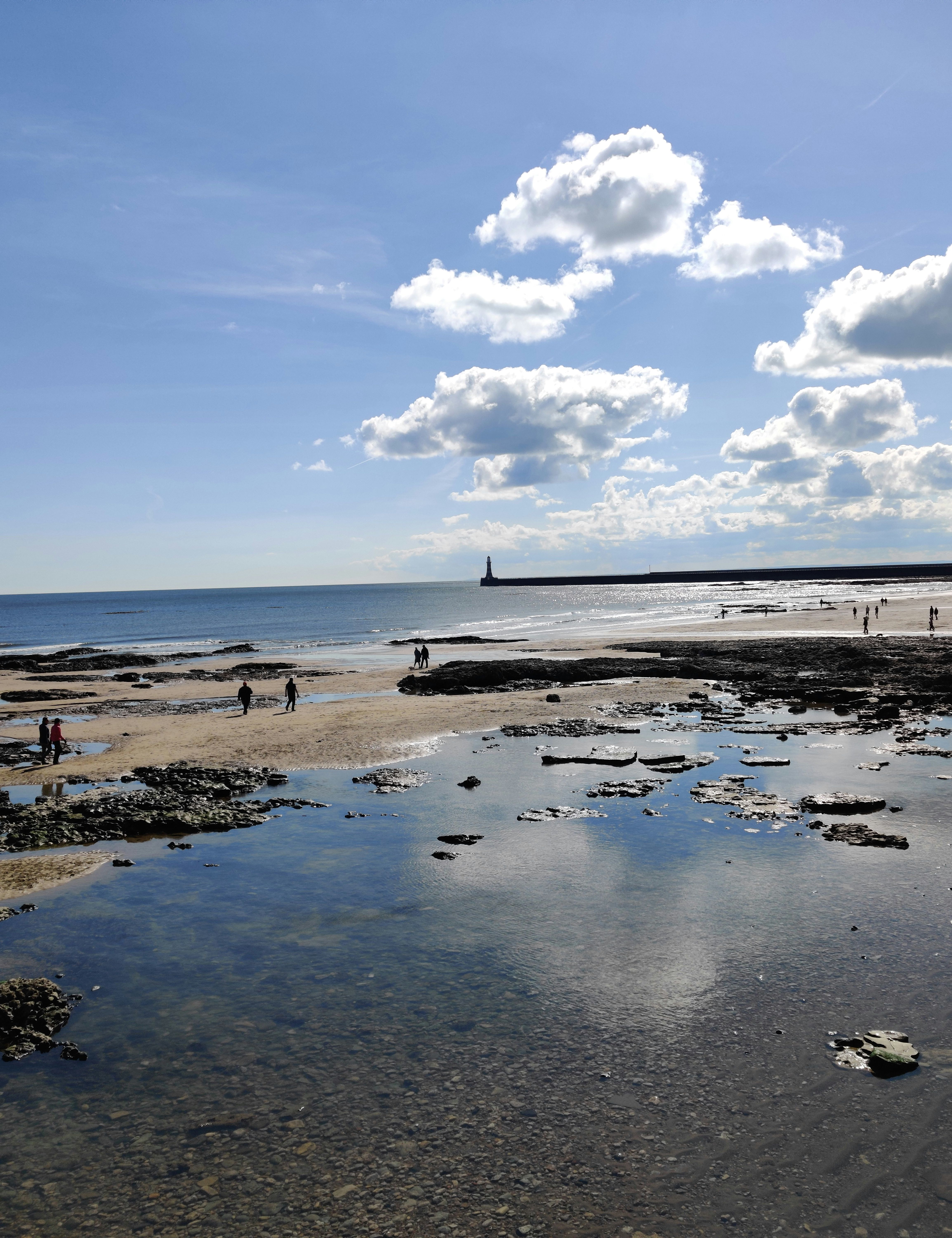 People walking along a rocky beach with shallow tide pools and a lighthouse in the distance under a blue sky with scattered clouds.