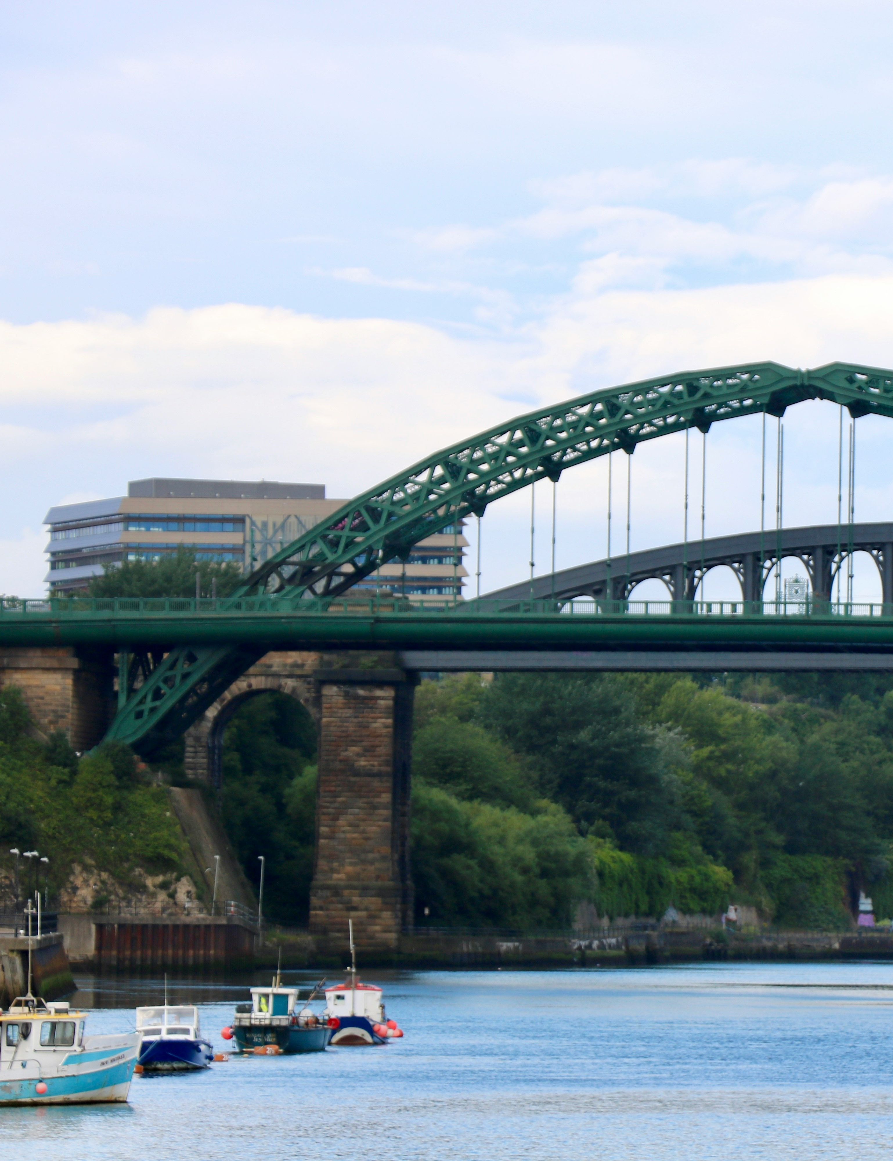 Green arched bridge spanning a river with boats below and trees and buildings in the background