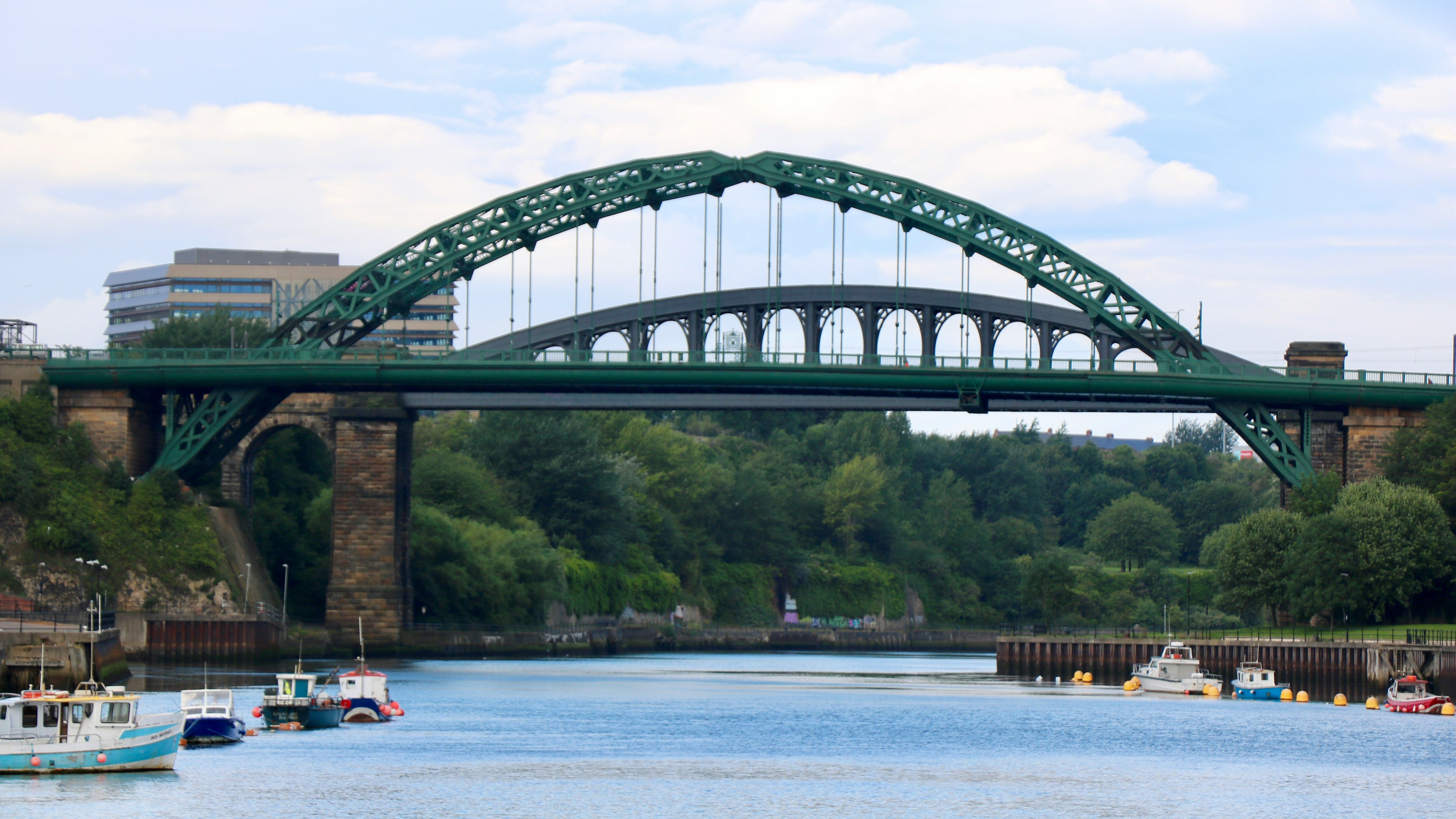 Green arched bridge spanning a river with boats below and trees and buildings in the background