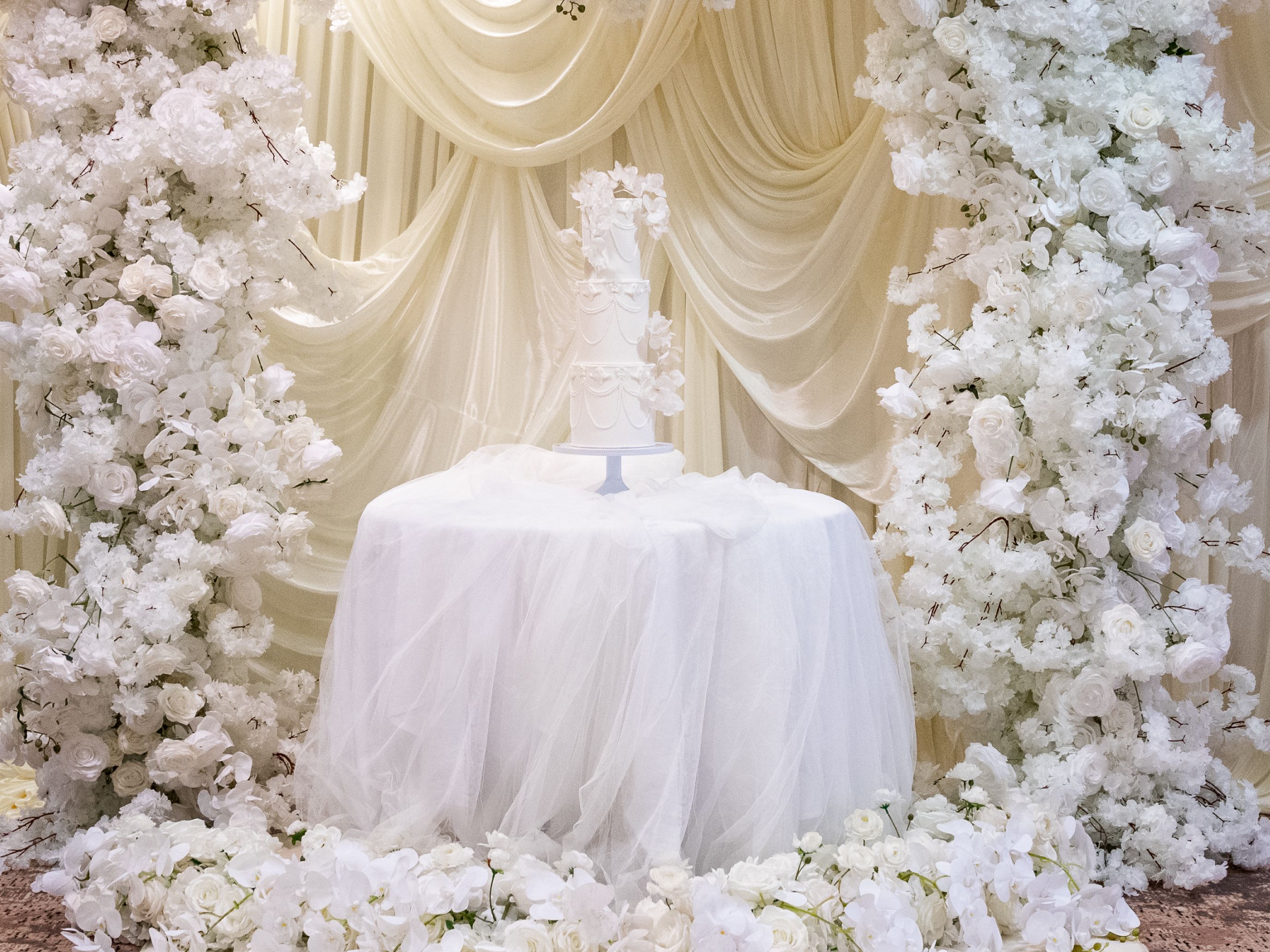 Wedding cake displayed under a floral arch with white flowers and draped fabric backdrop