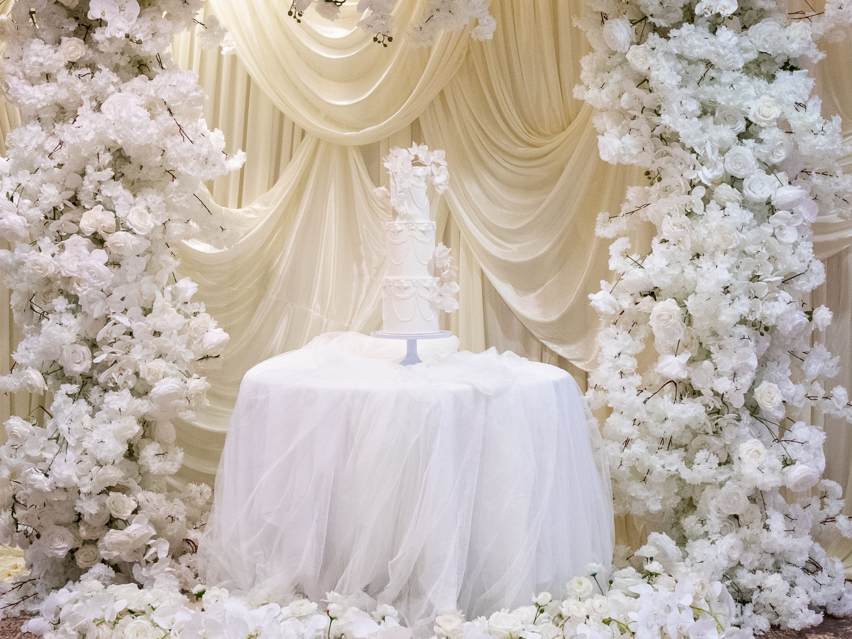 Wedding cake displayed under a floral arch with white flowers and draped fabric backdrop
