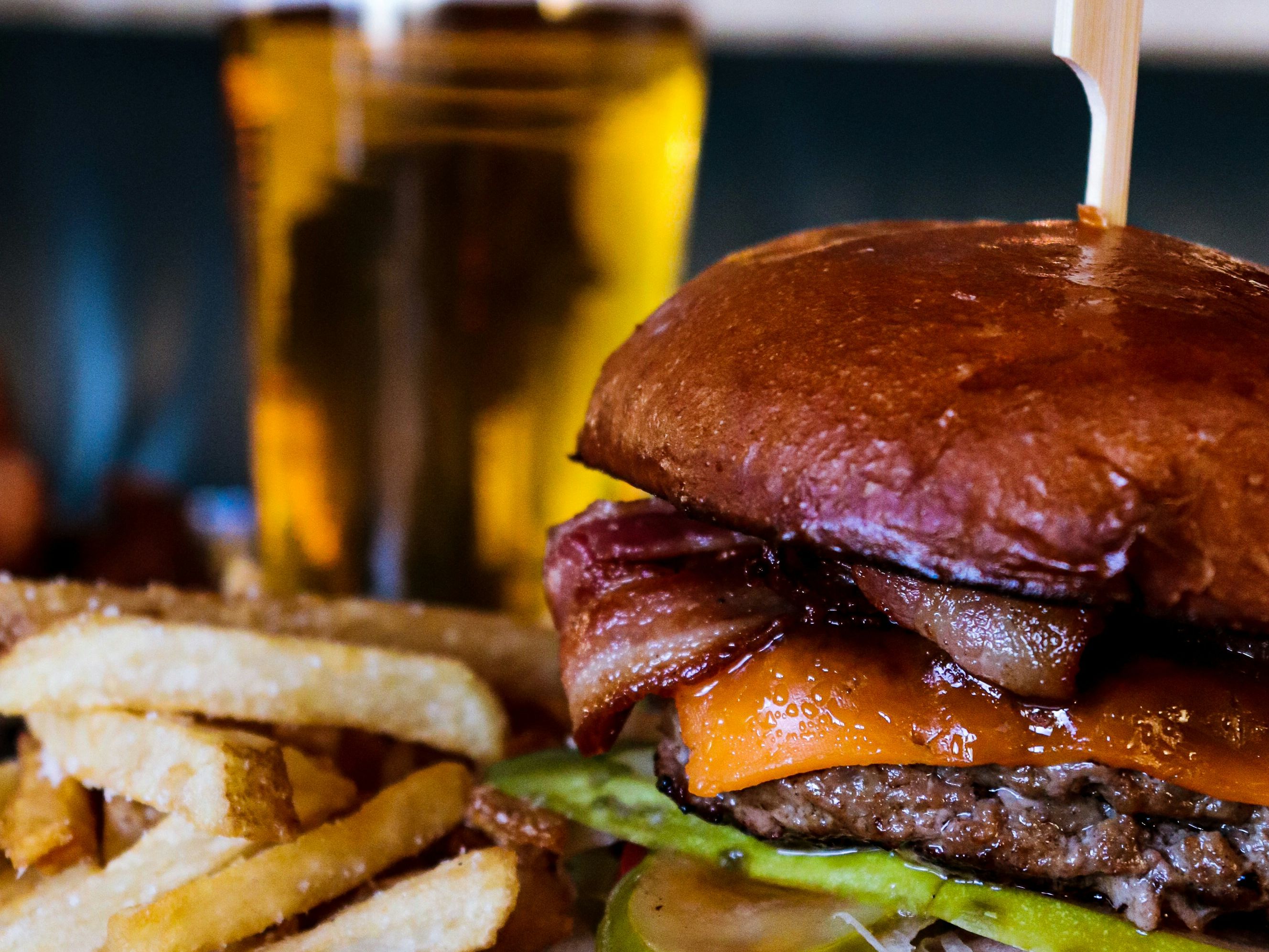 Close-up of a cheeseburger with bacon, lettuce, tomato, pickles, and a side of French fries, with a glass of beer in the background.