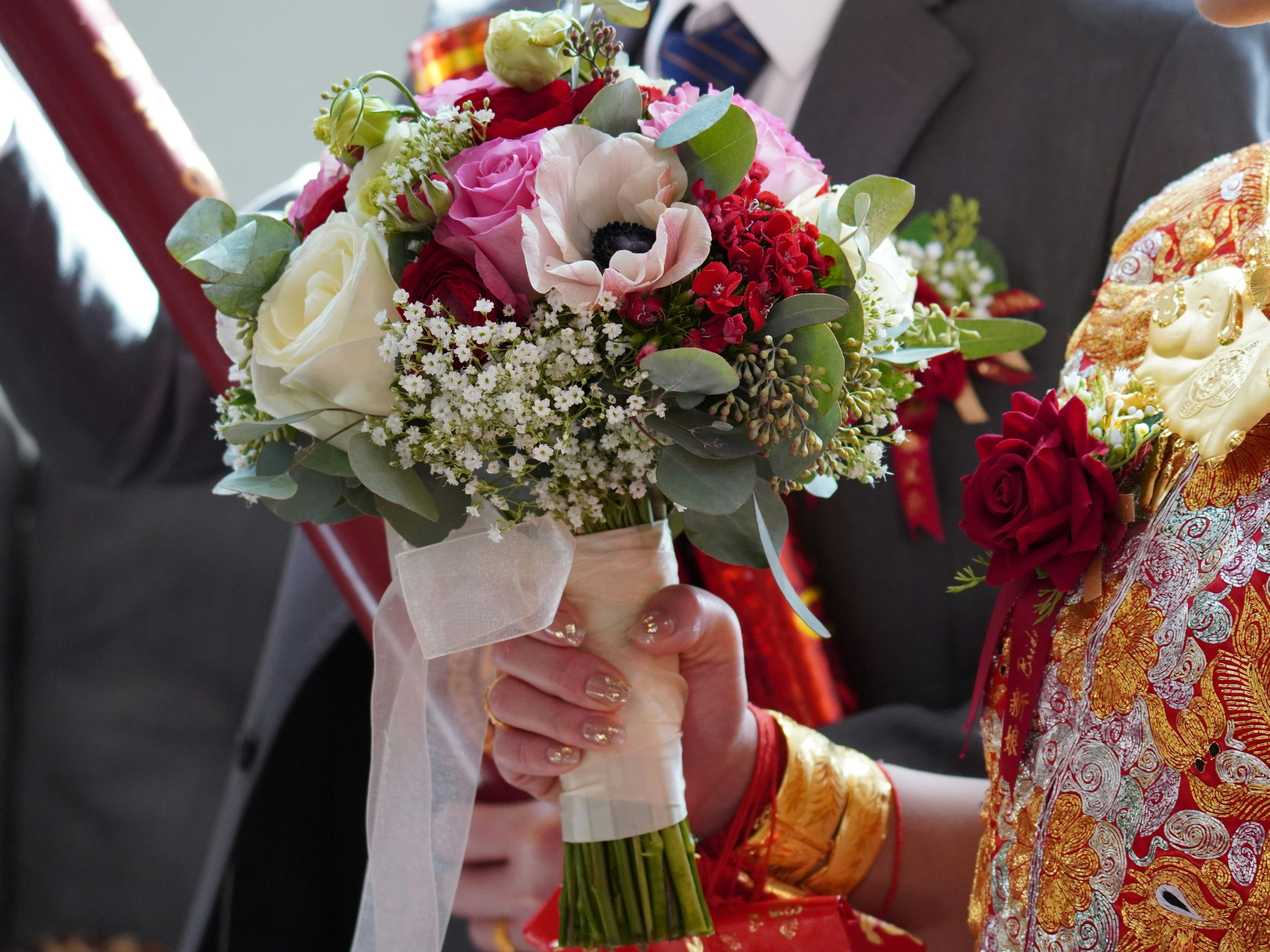 Person in ornate traditional clothing holding a vibrant wedding bouquet with white, pink, and red flowers.