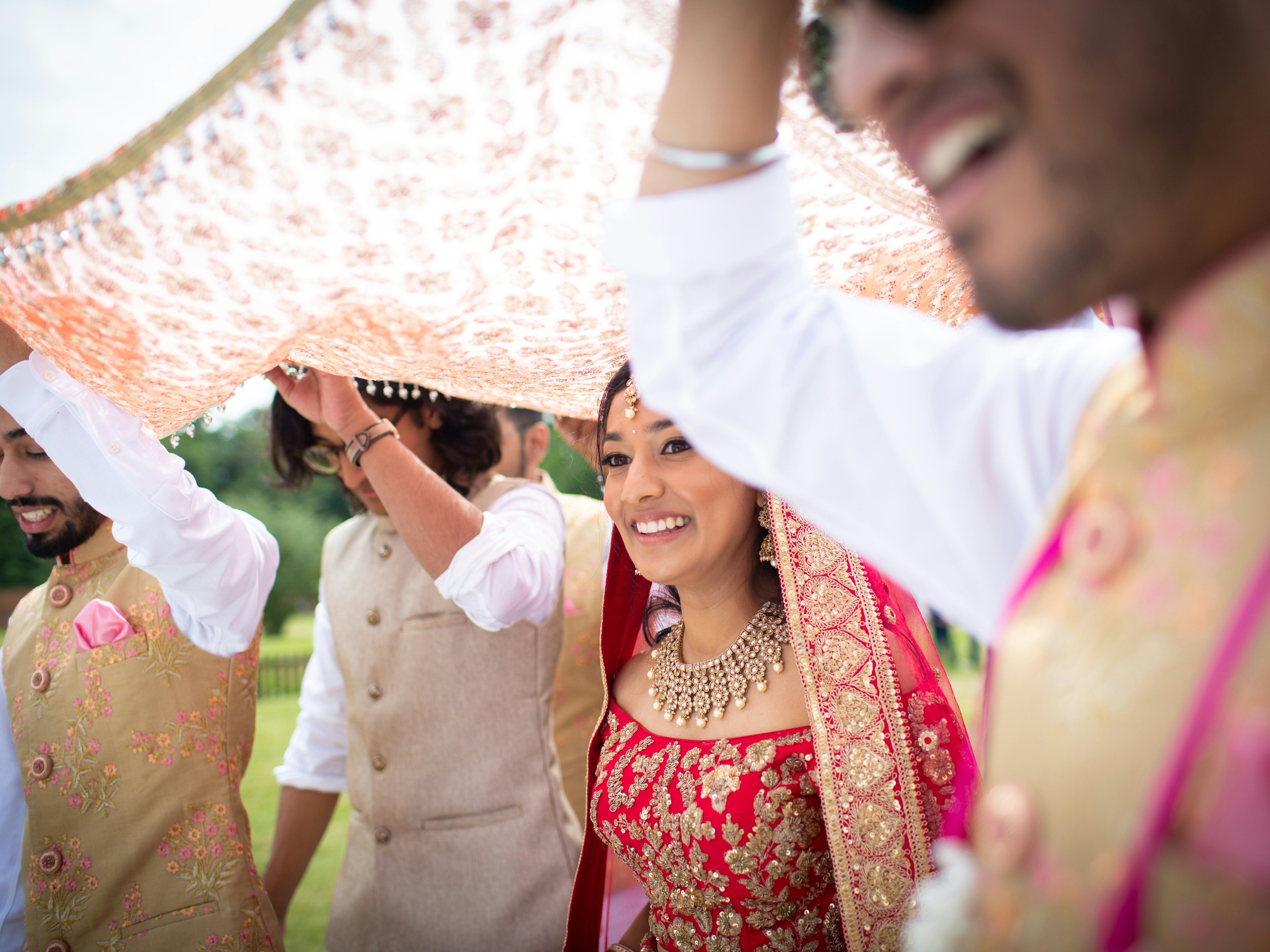 Bride in traditional Indian attire being escorted under a decorative cloth during a wedding ceremony