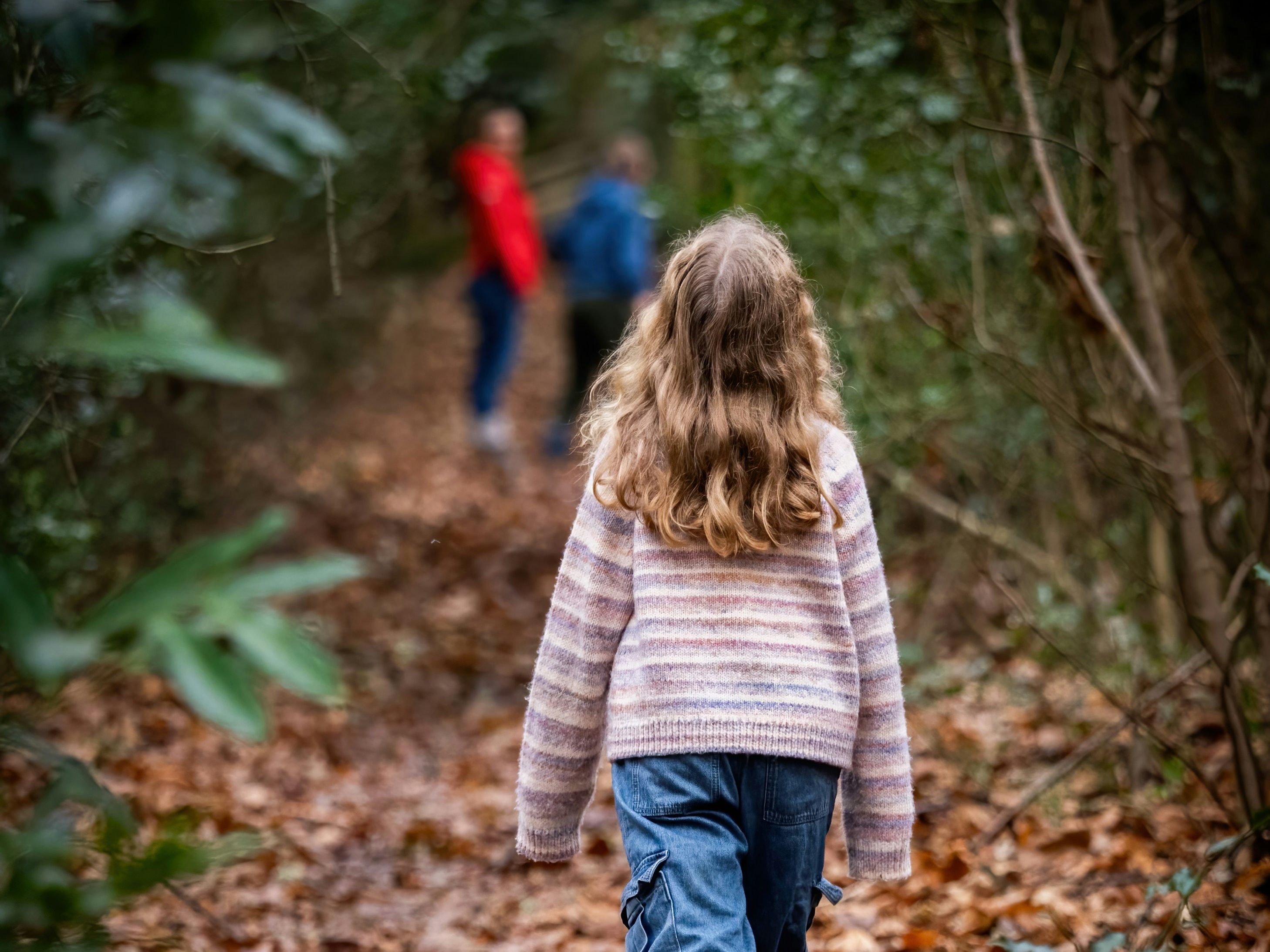 Child walking on a leaf-covered path in the woods with two blurred figures in the background.