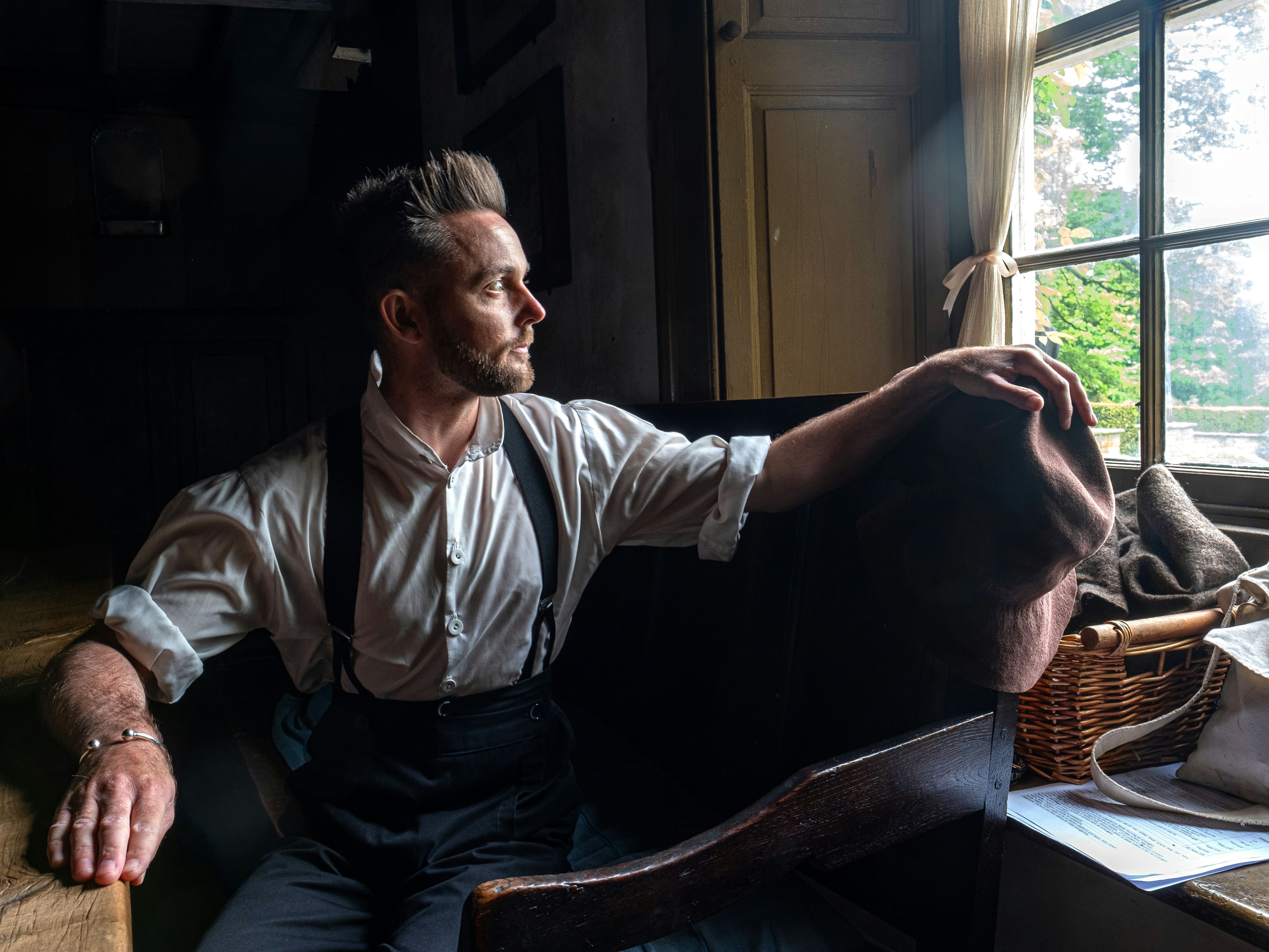 Man in suspenders sitting by a window, looking outside with a contemplative expression.